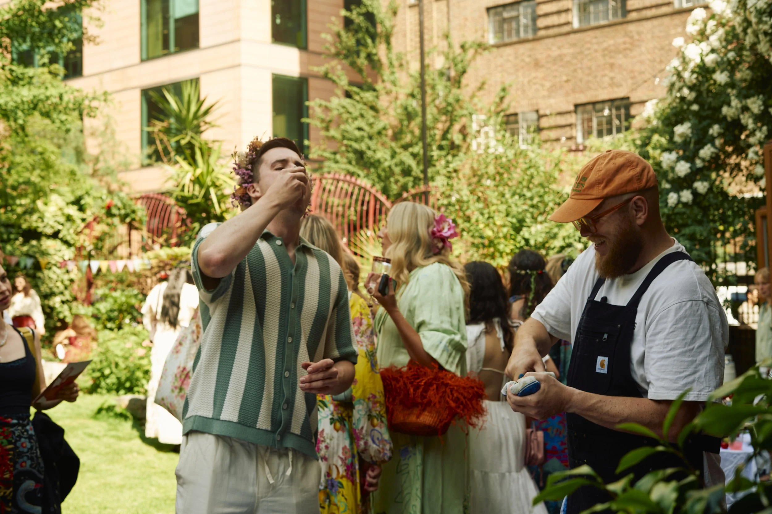 People at a garden party, some wearing floral crowns, enjoying drinks and socializing among greenery and urban buildings.