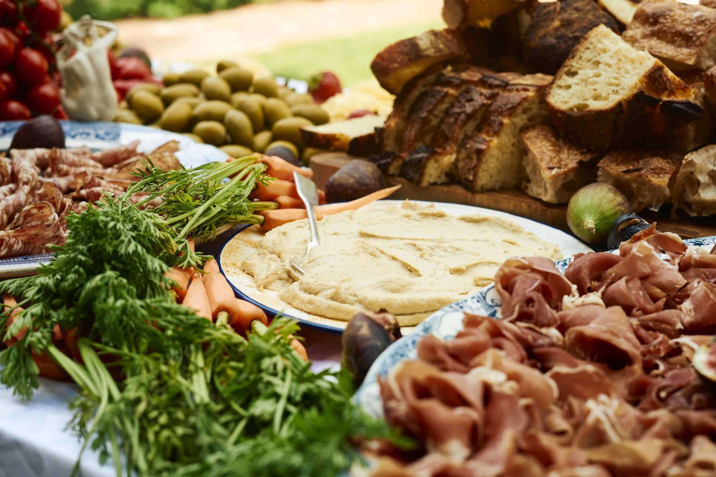 A spread of Mediterranean food including hummus, fresh vegetables, pita bread, olives, and cured meats on a table.