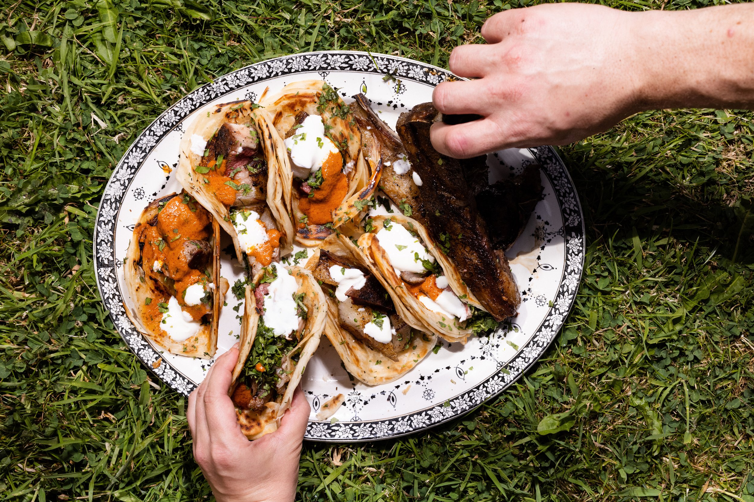 Plate of tacos with meat, topped with white sauce, orange sauce, and chopped herbs, placed on grass, with two hands reaching for the food.