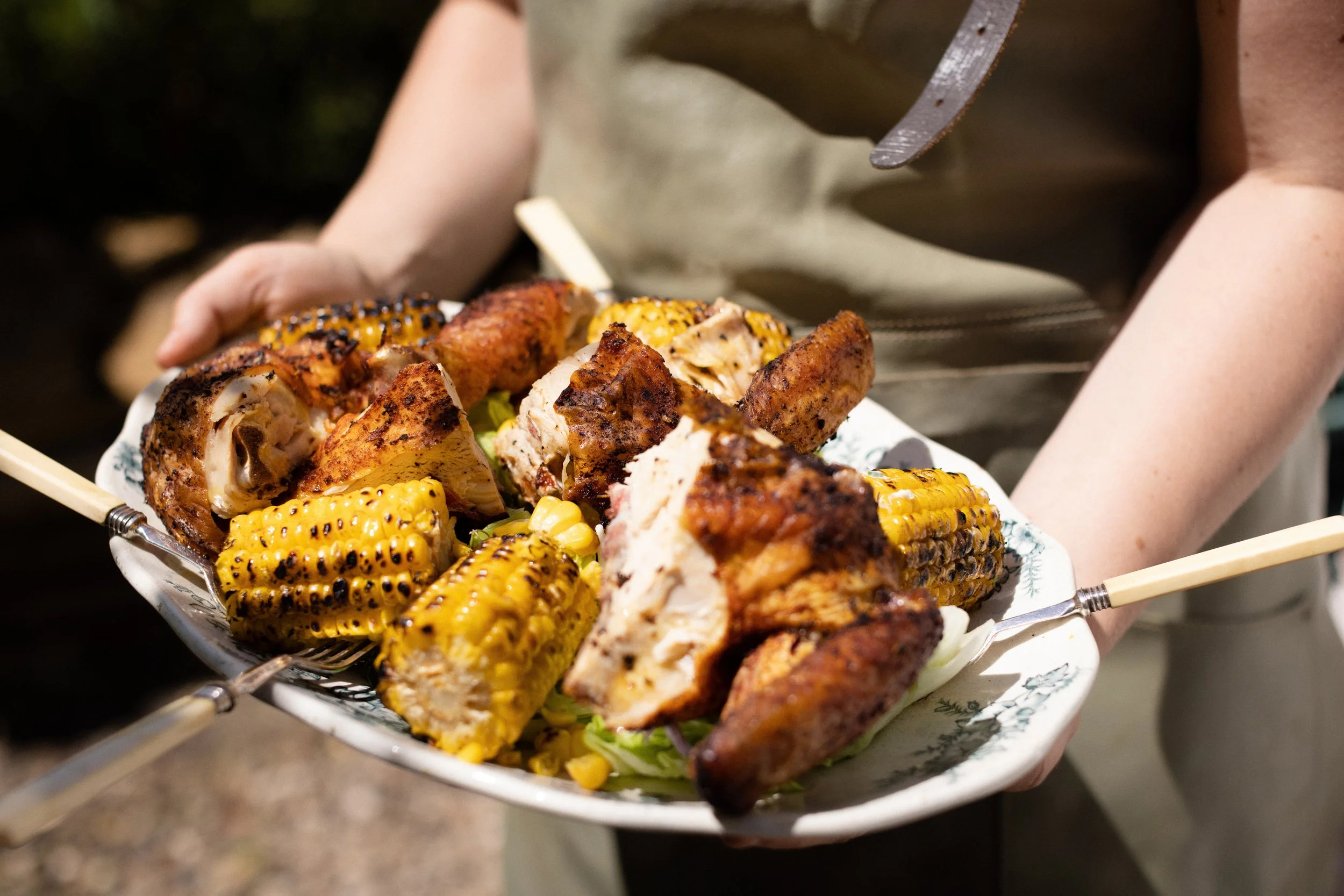 Person holding a plate of grilled chicken thighs and corn on the cob.