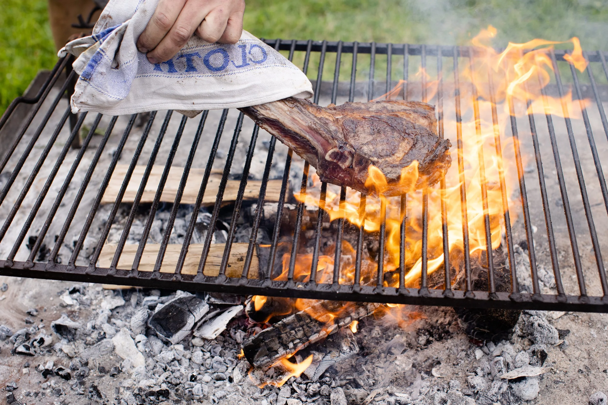 A person grilling a large piece of meat over an open flame on a barbecue grill outdoors.