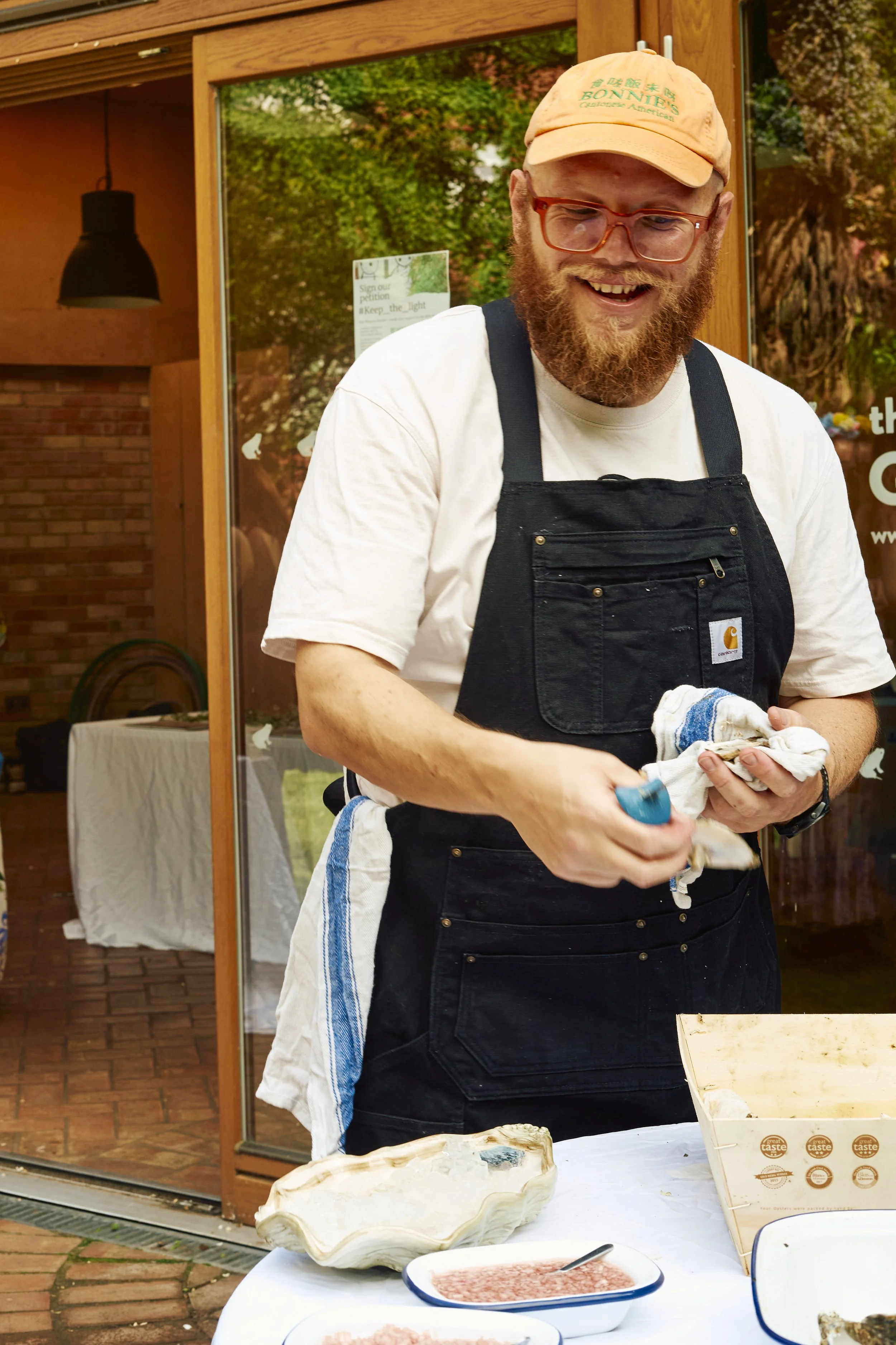 A man with red hair, a beard, glasses, wearing a beige cap, a white t-shirt, and a black apron, smiling while preparing food outside a building.