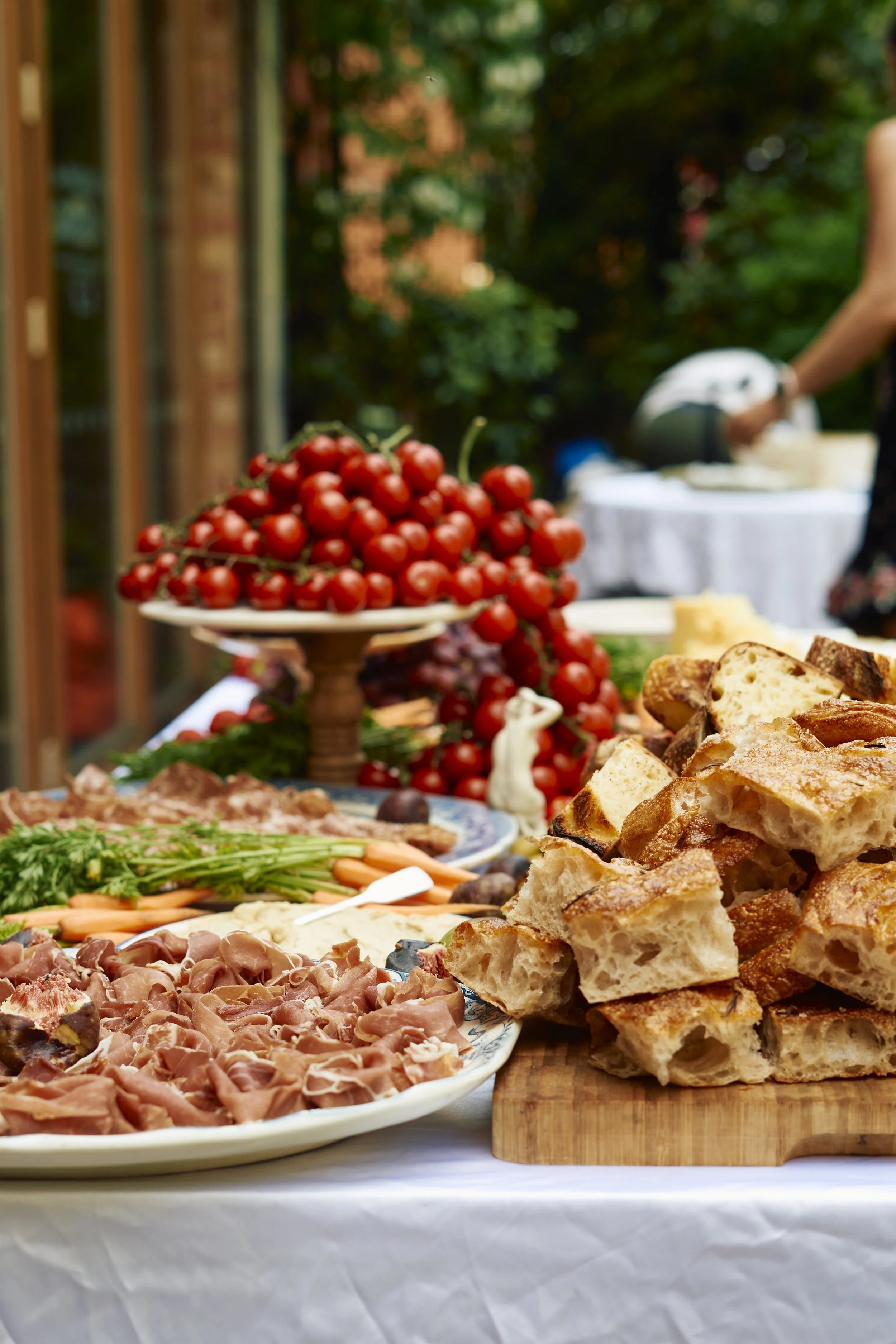 A table set outdoors with various foods including cherry tomatoes, sliced bread, prosciutto, vegetables, and cheeses, indicating a buffet or picnic setting.