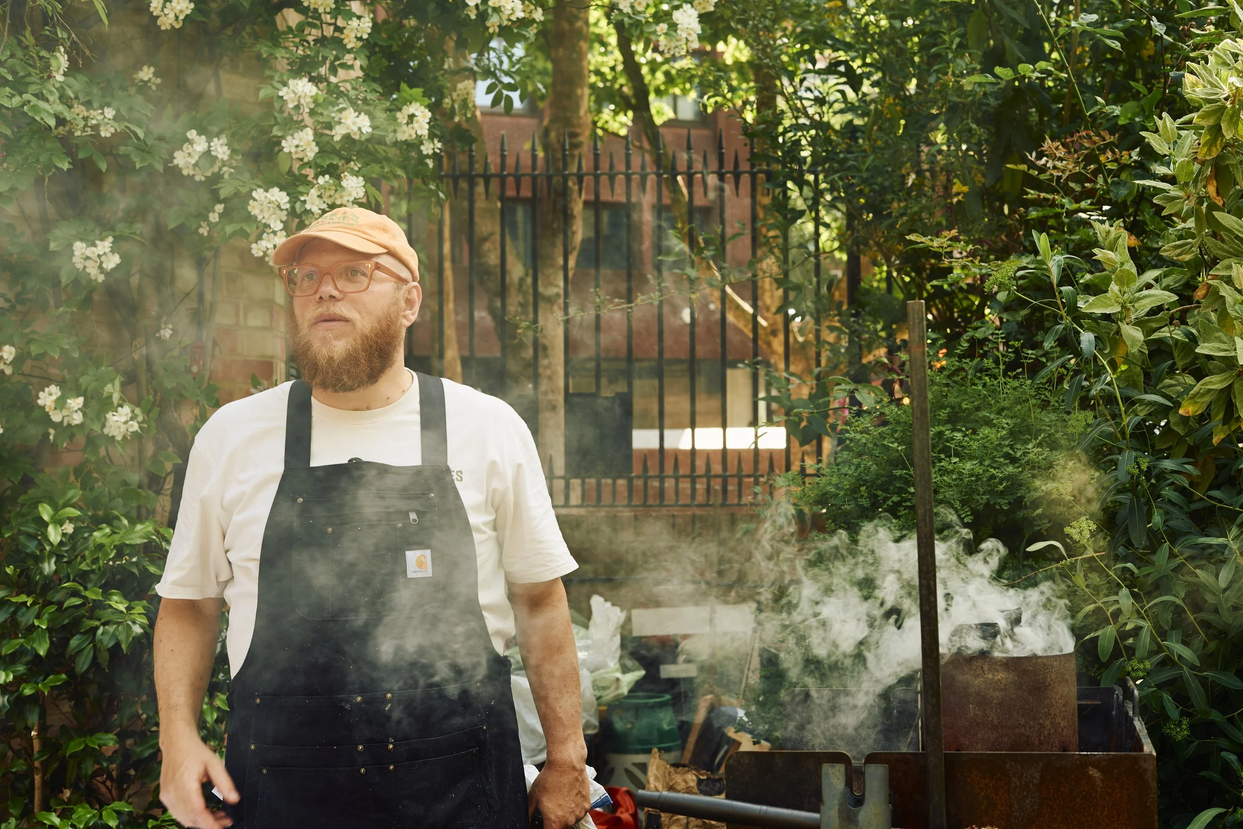 A man with glasses and a beard wearing a beige cap and white T-shirt, standing outdoors in a lush garden with flowering plants and greenery, next to a barbecue grill emitting smoke.