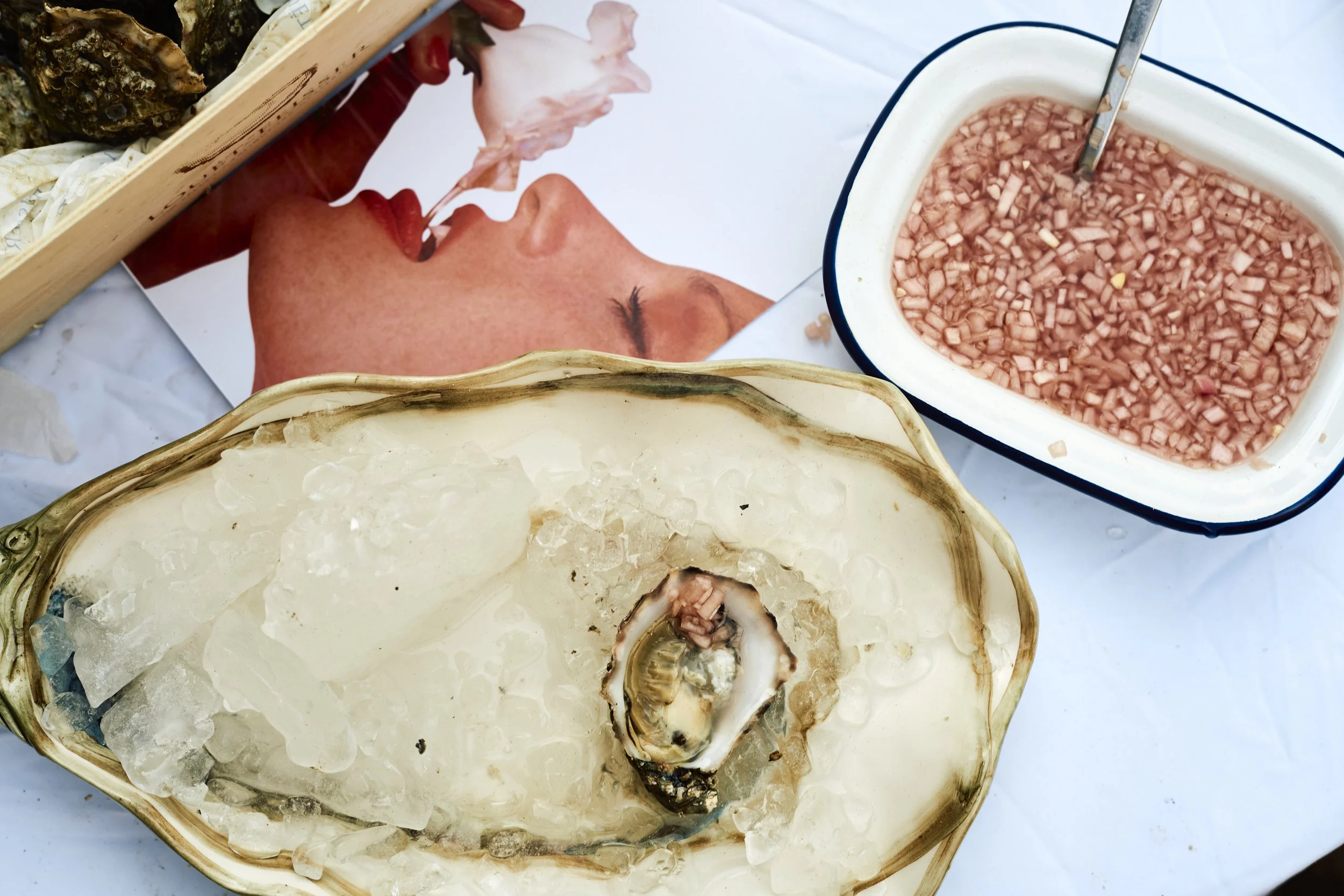 A basket with ice, a clam shell with a clam, and a container of chopped purple onions, with a magazine showing a woman with red hair and a shell on the page.