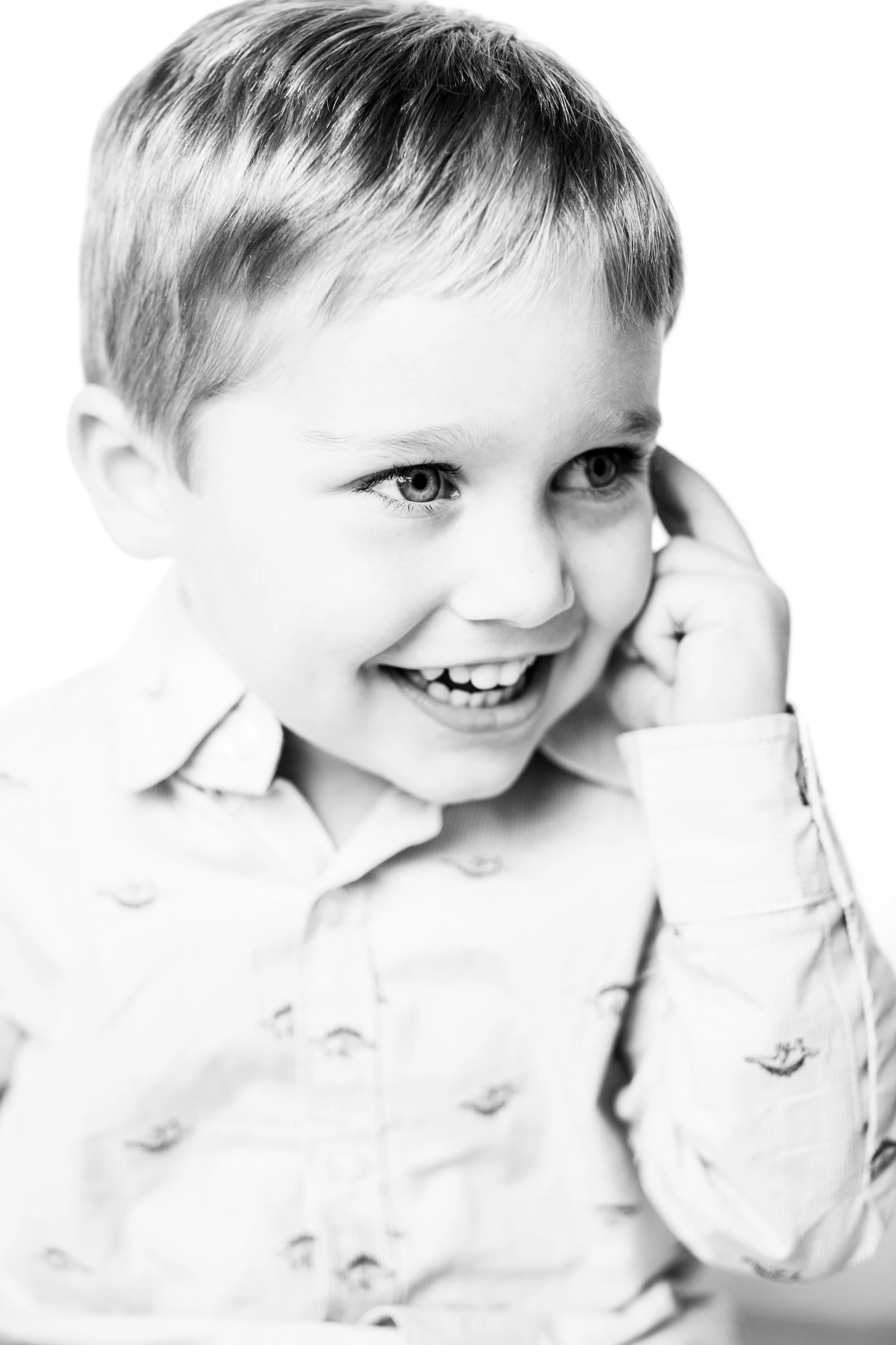 A young boy with short hair, smiling and holding his ear with his hand, wearing a light-colored button-up shirt with small bird prints, in black and white.