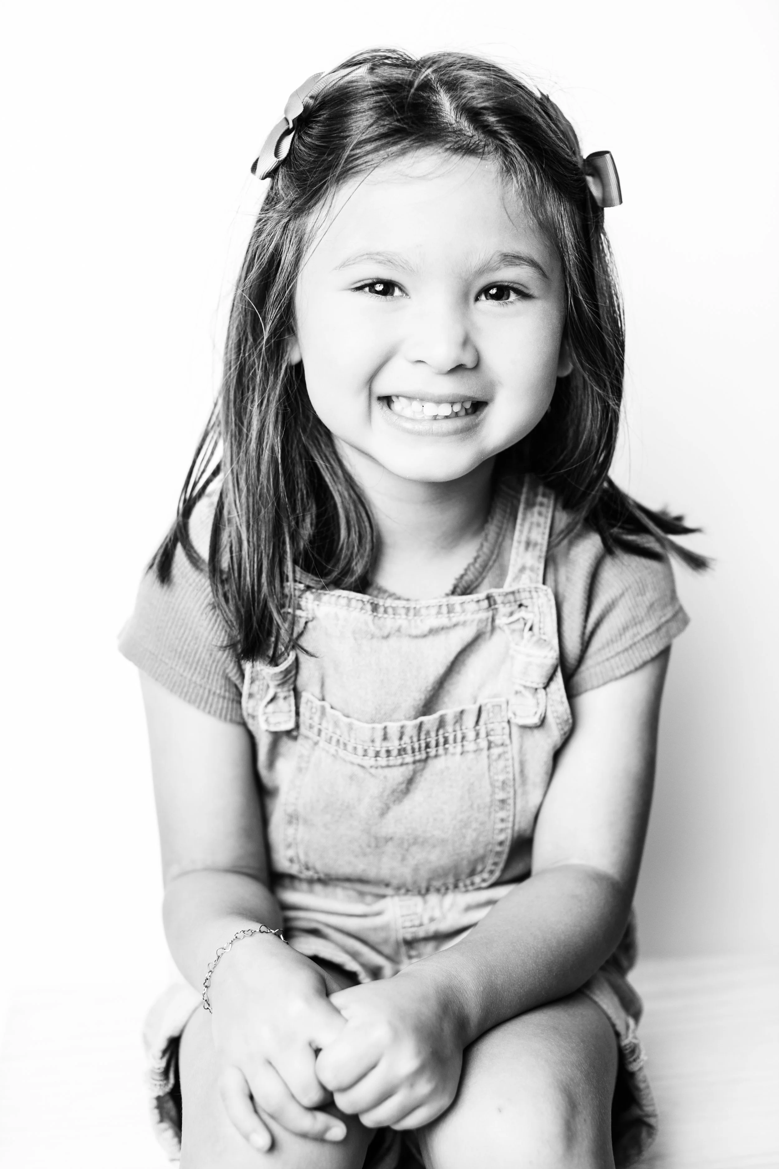 A young girl with long hair, wearing a striped shirt and overalls, smiling at the camera with her hands clasped on her lap, in a black-and-white photo.