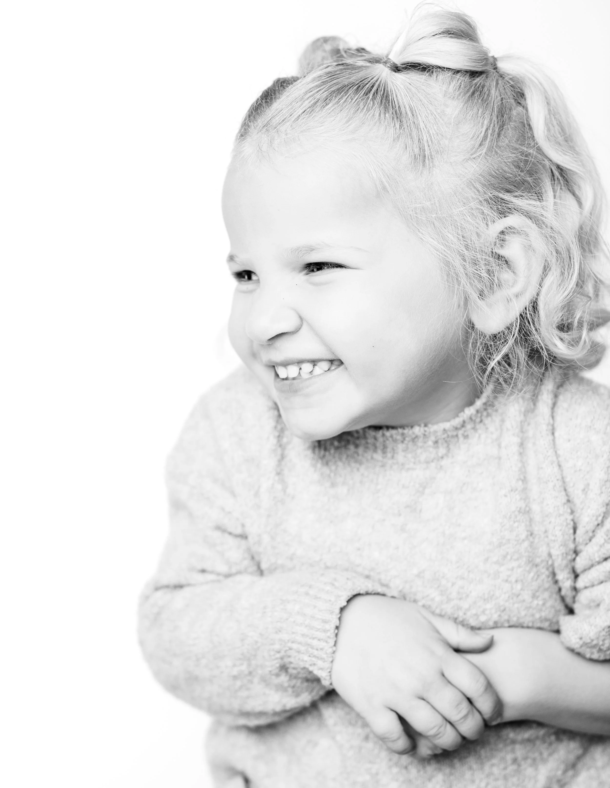 A young girl with curly hair smiling and looking to her right, wearing a light-colored sweater.