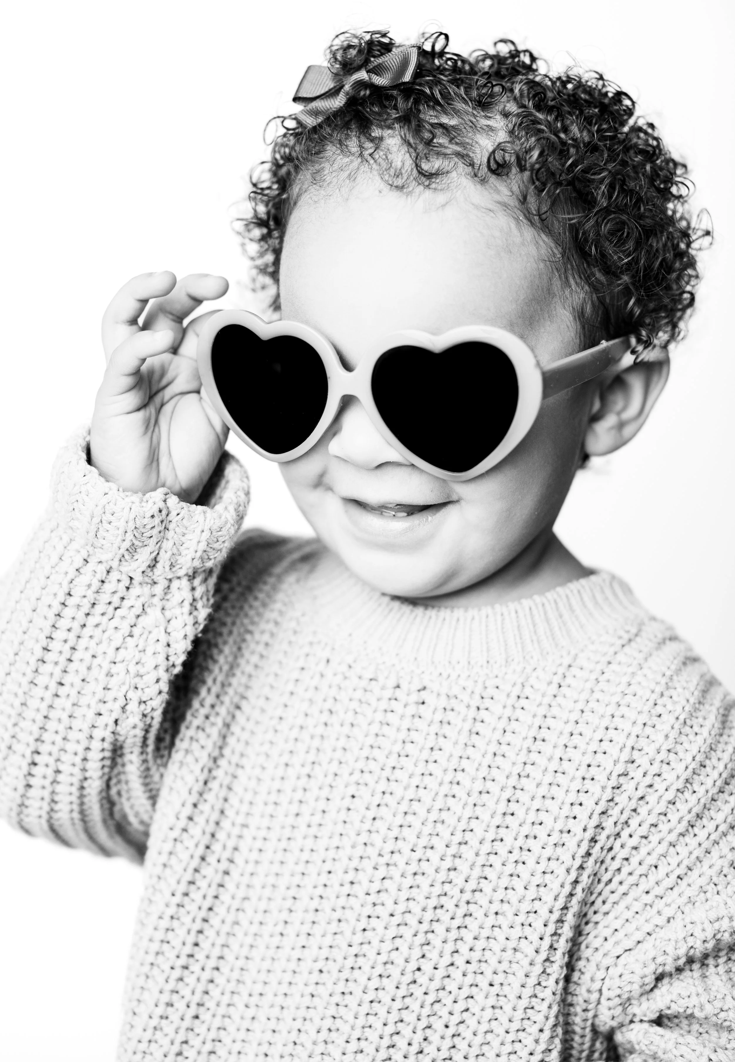 A young girl with curly hair, wearing a bow and sunglasses, smiling and looking playful.