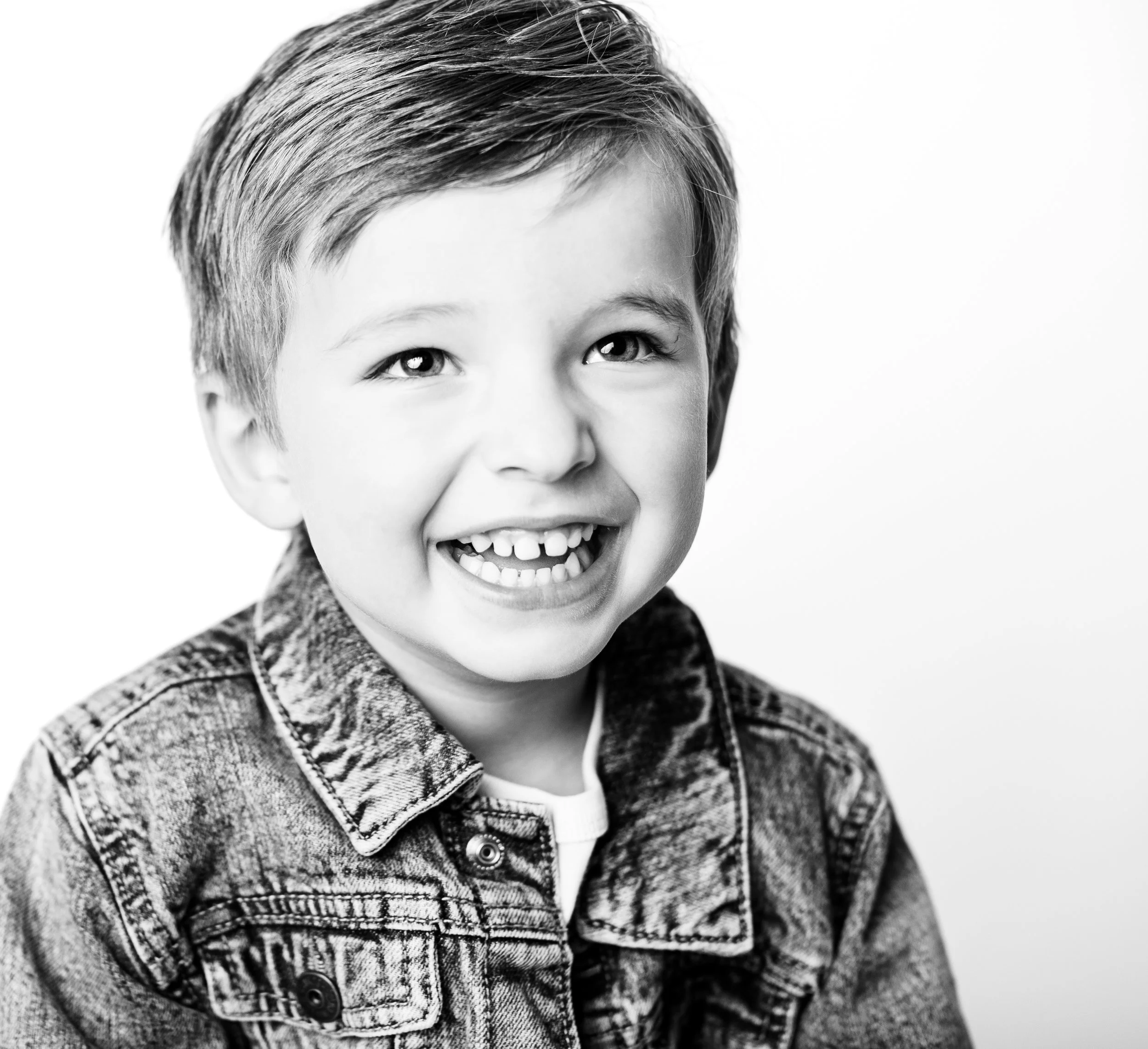 Black and white photo of a smiling young boy with short hair, wearing a denim jacket.