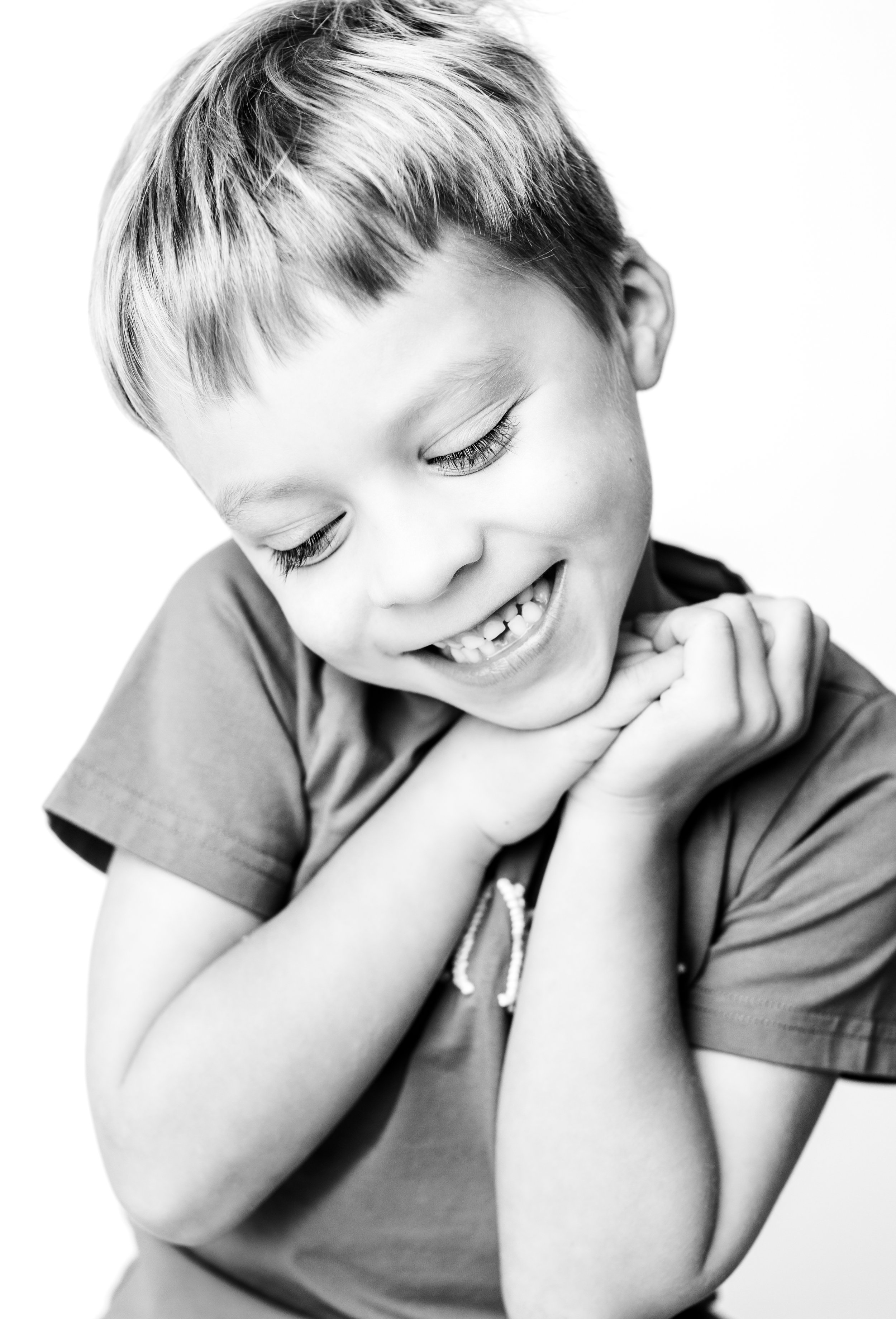 Black and white photo of a smiling young boy with closed eyes, holding his hands together near his chin, wearing a t-shirt.