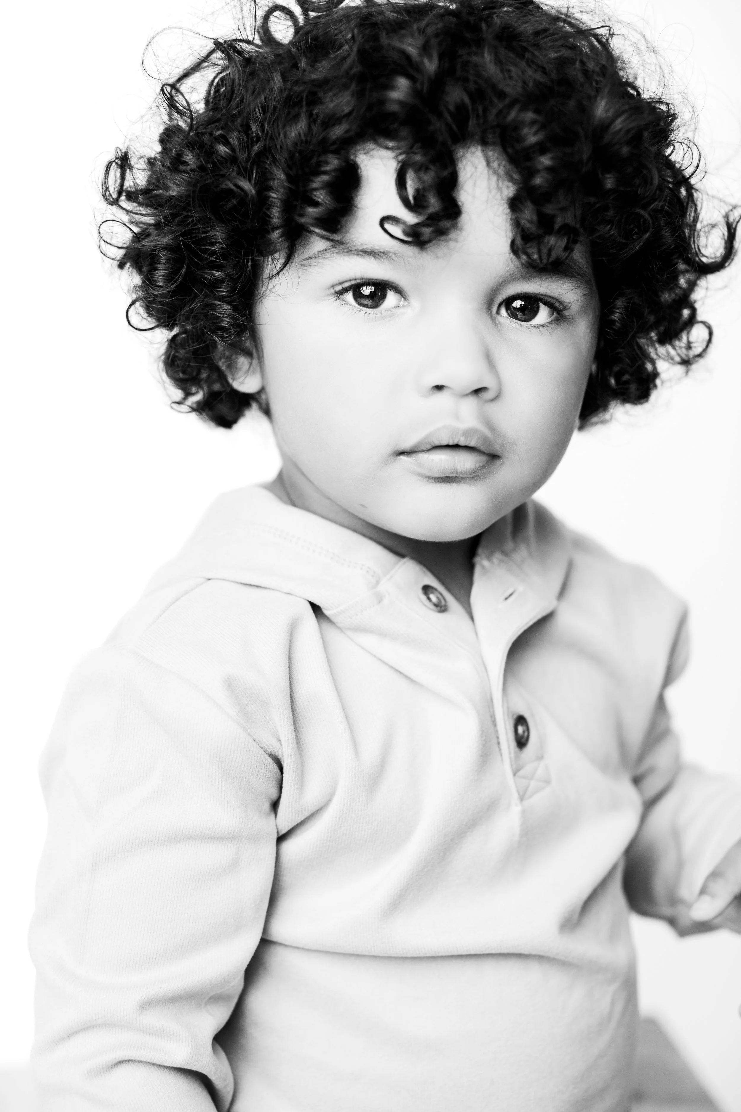 Black and white portrait of a young child with curly hair and serious expression, wearing a light-colored hoodie.