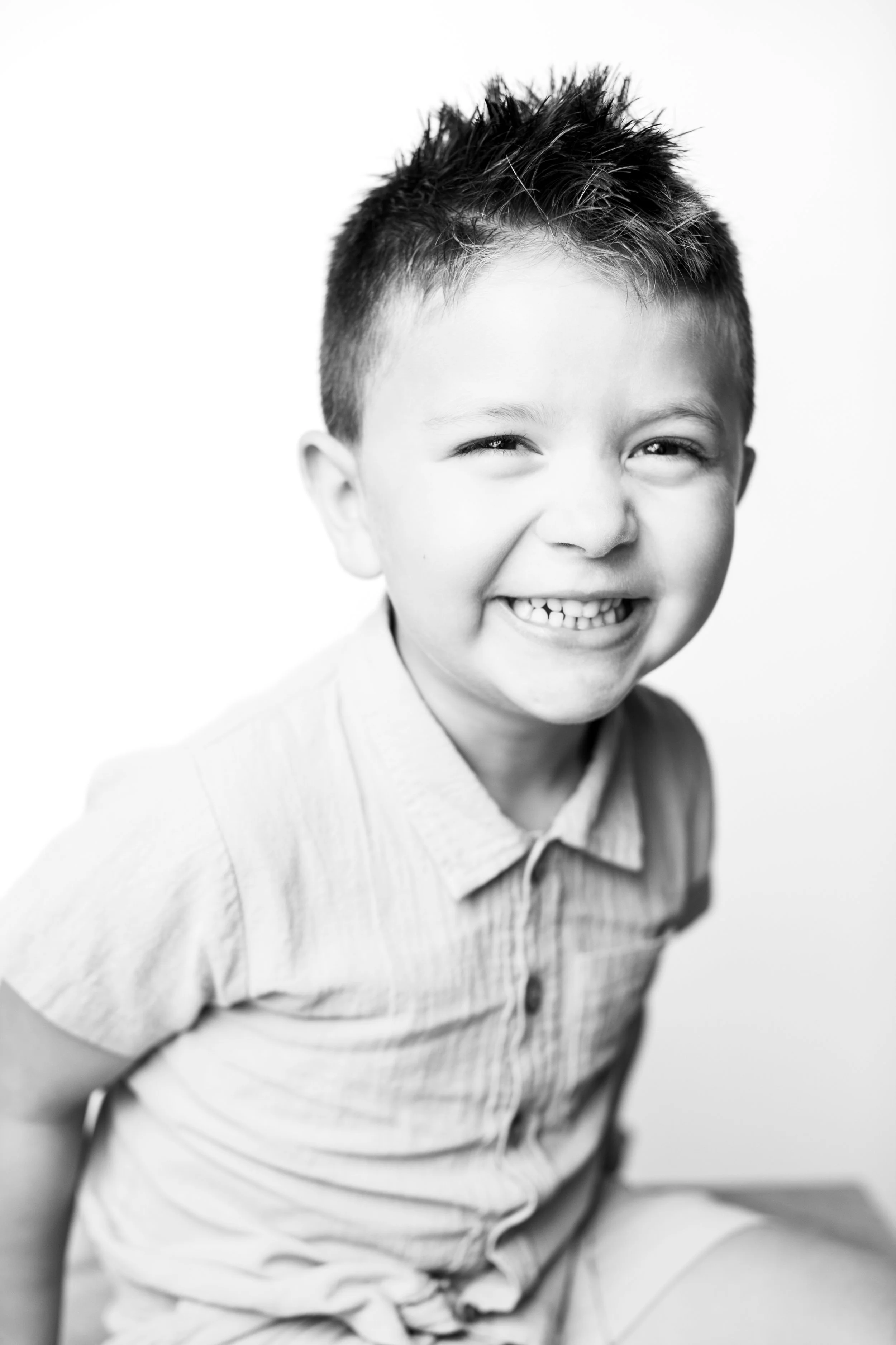A smiling young boy with spiky hair looks at the camera, wearing a vertically striped button-up shirt, in a black-and-white portrait.