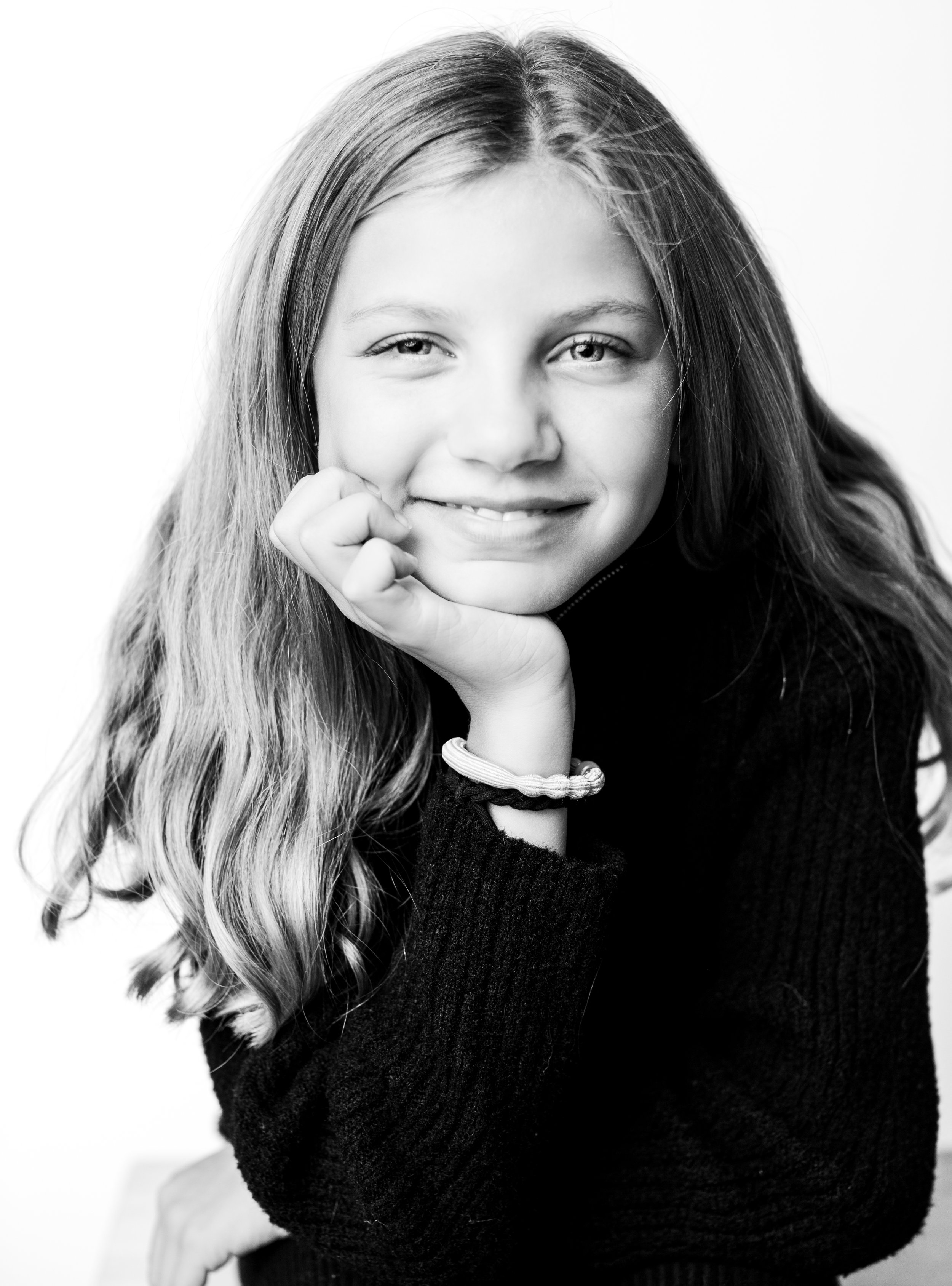 Black and white portrait of a young girl with long, wavy hair, smiling and resting her chin on her hand, wearing a dark sweater.