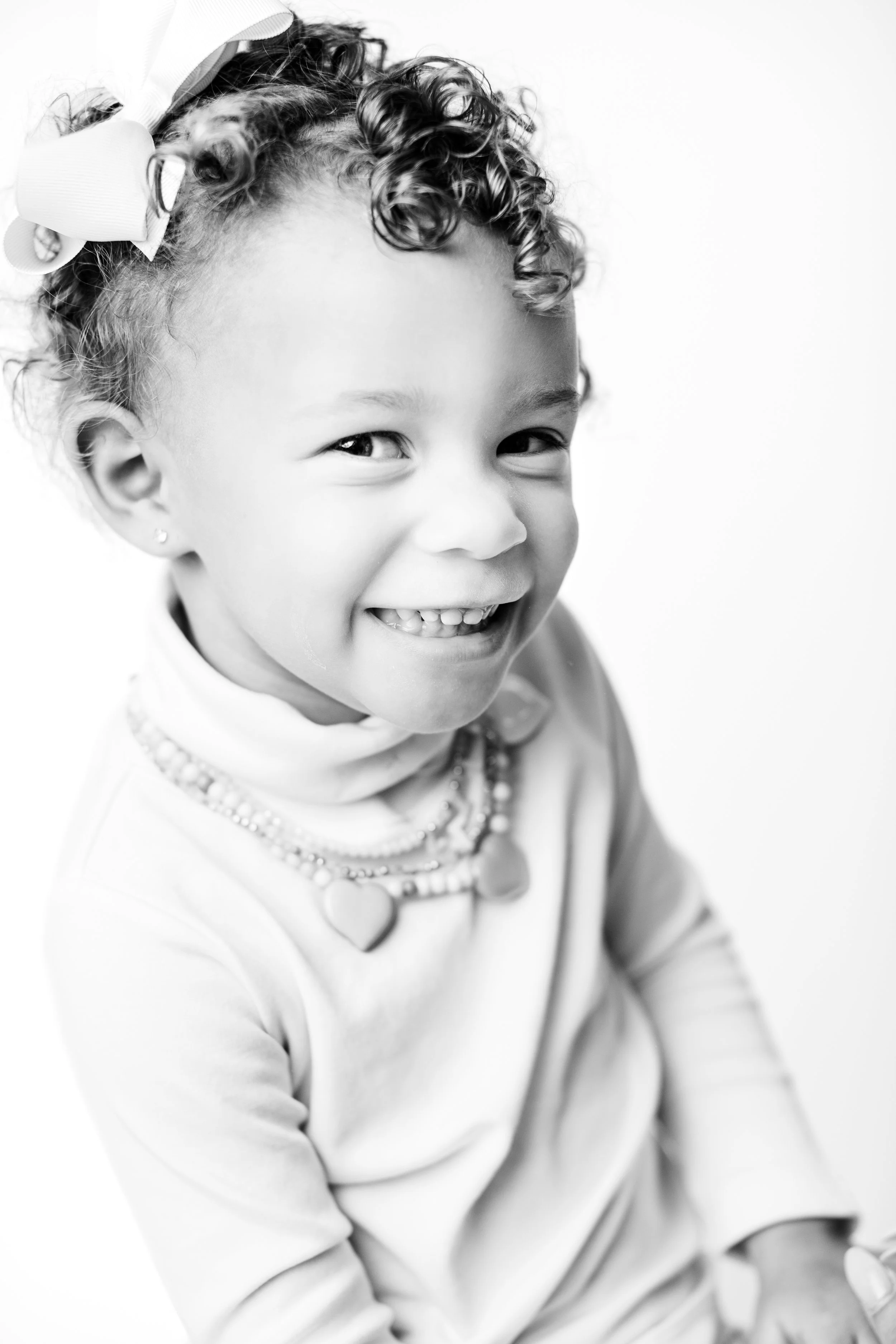 Smiling young girl with curly hair, wearing a bow, jewelry, and a long-sleeved top, against a plain background.
