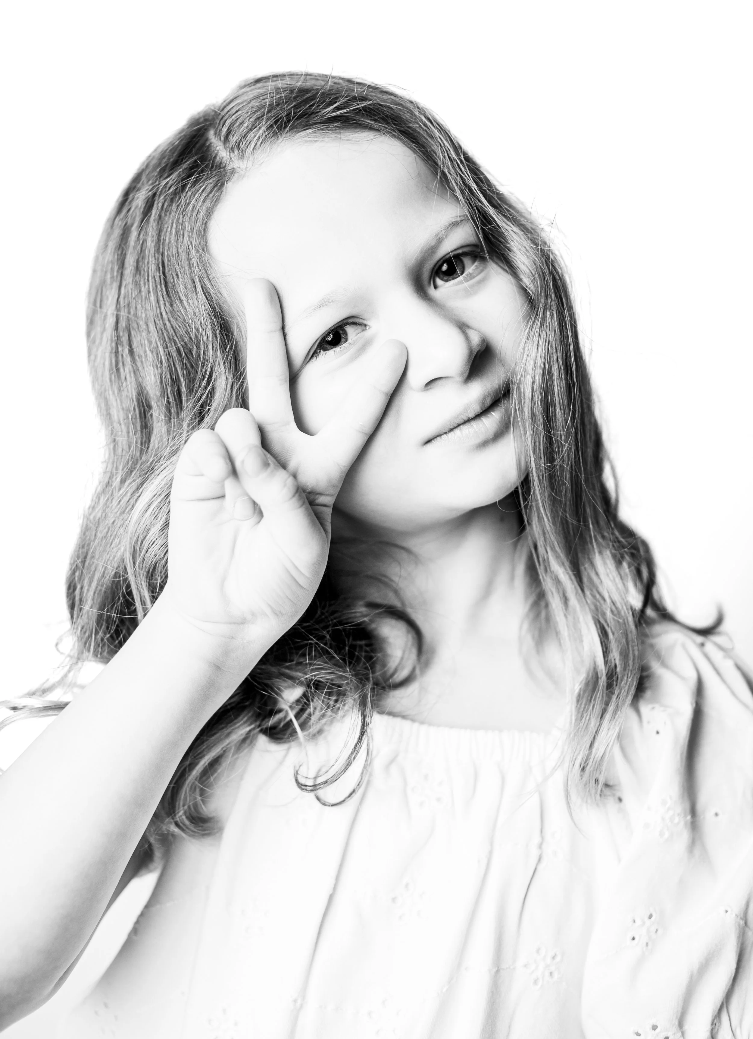 A black and white photograph of a young girl with wavy hair making a peace sign near her eye.