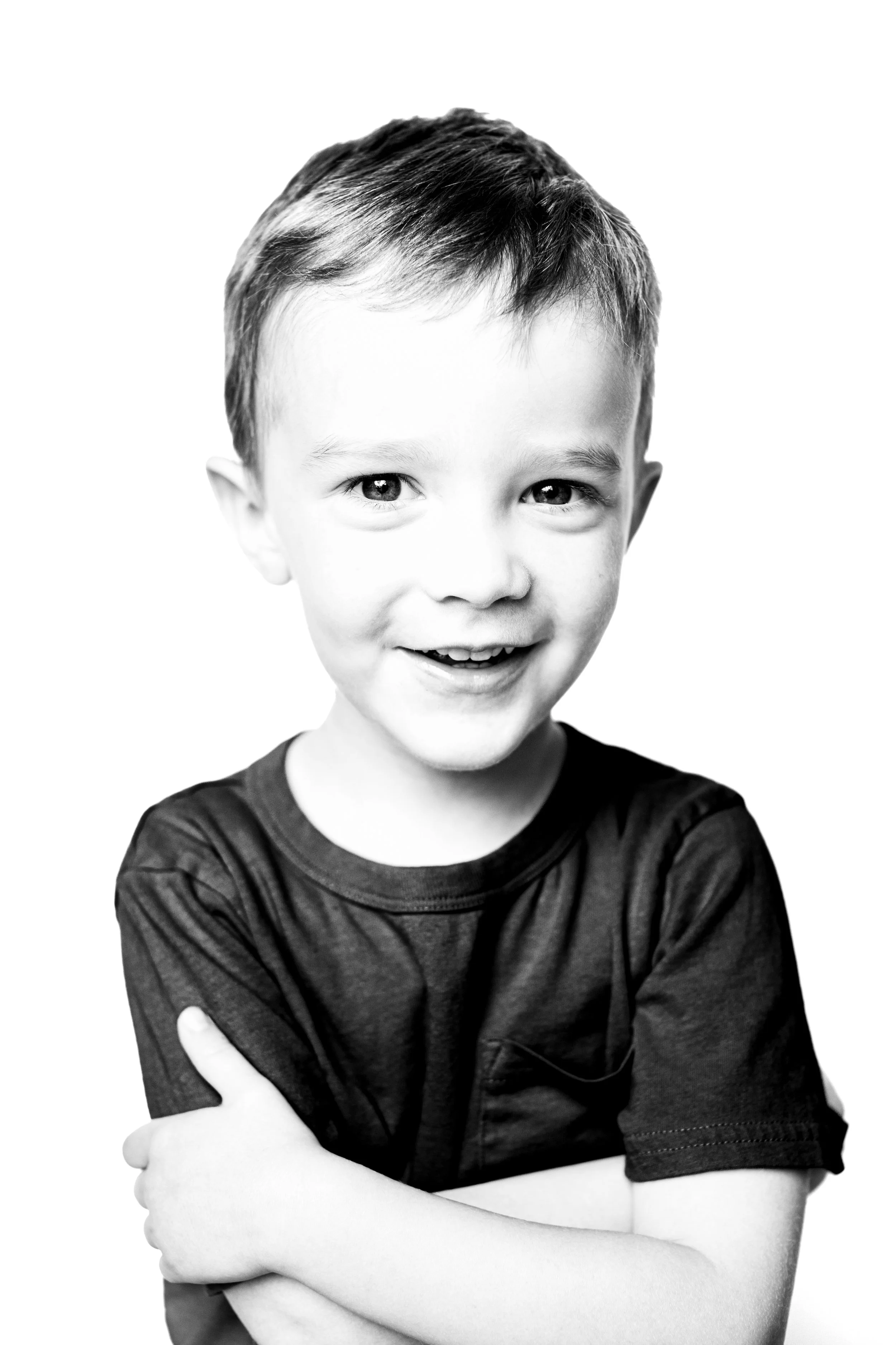 A smiling young boy with short hair, arms crossed, wearing a dark t-shirt, black and white portrait.