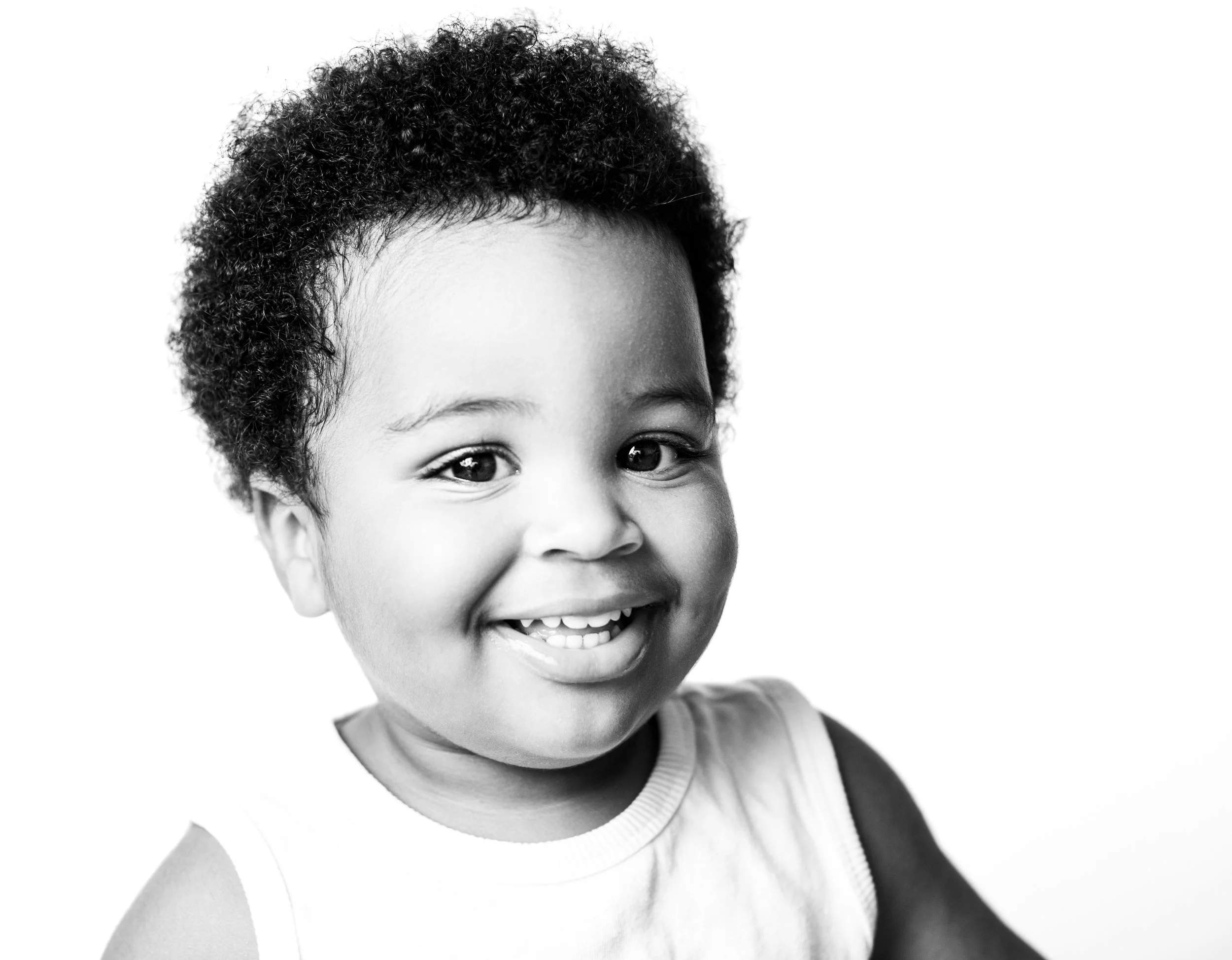 A smiling young child with curly hair in a sleeveless shirt, black-and-white photo, facing the camera.