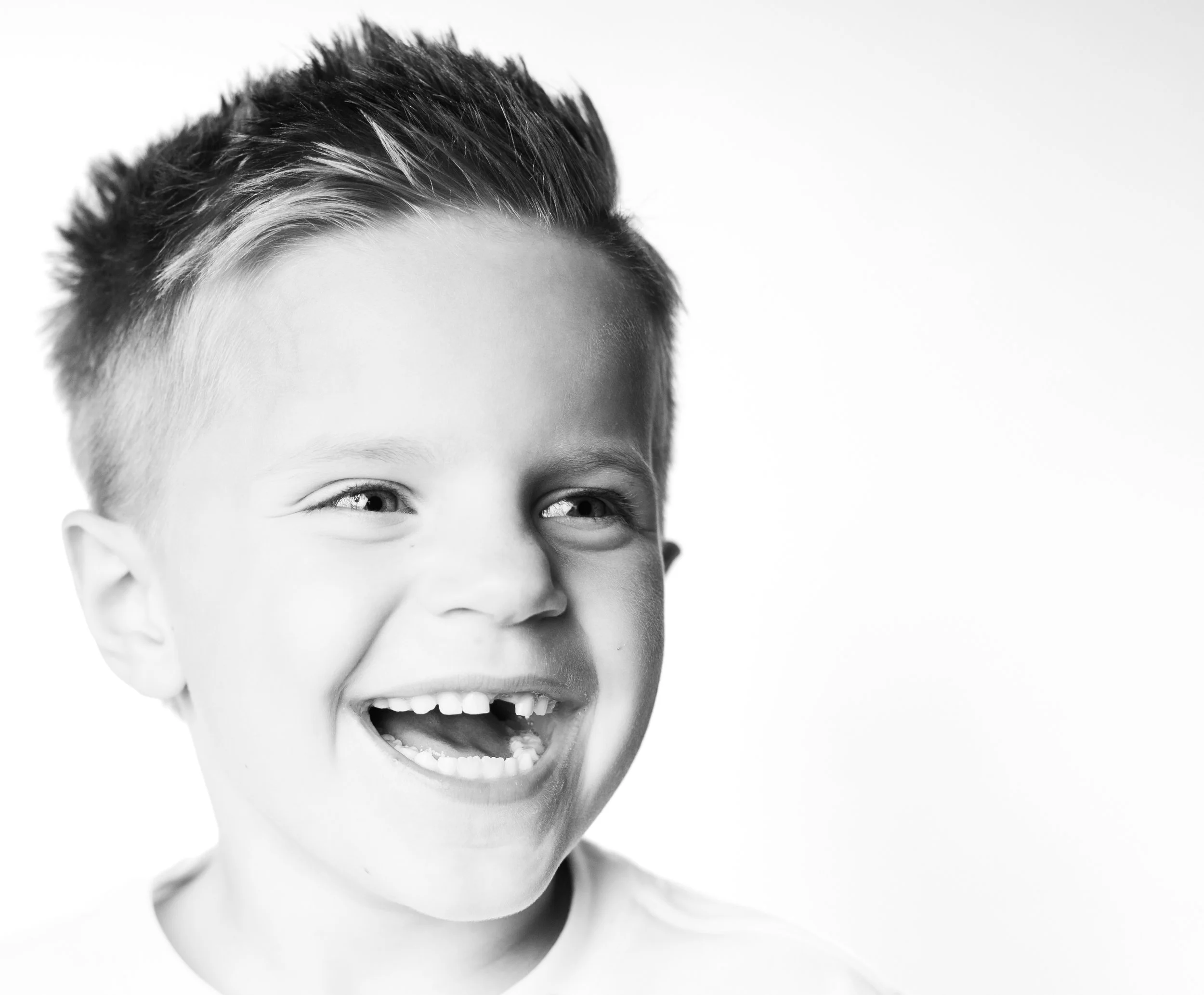 Black and white close-up of a smiling young boy with short, styled hair, showing missing front teeth, against a plain background.