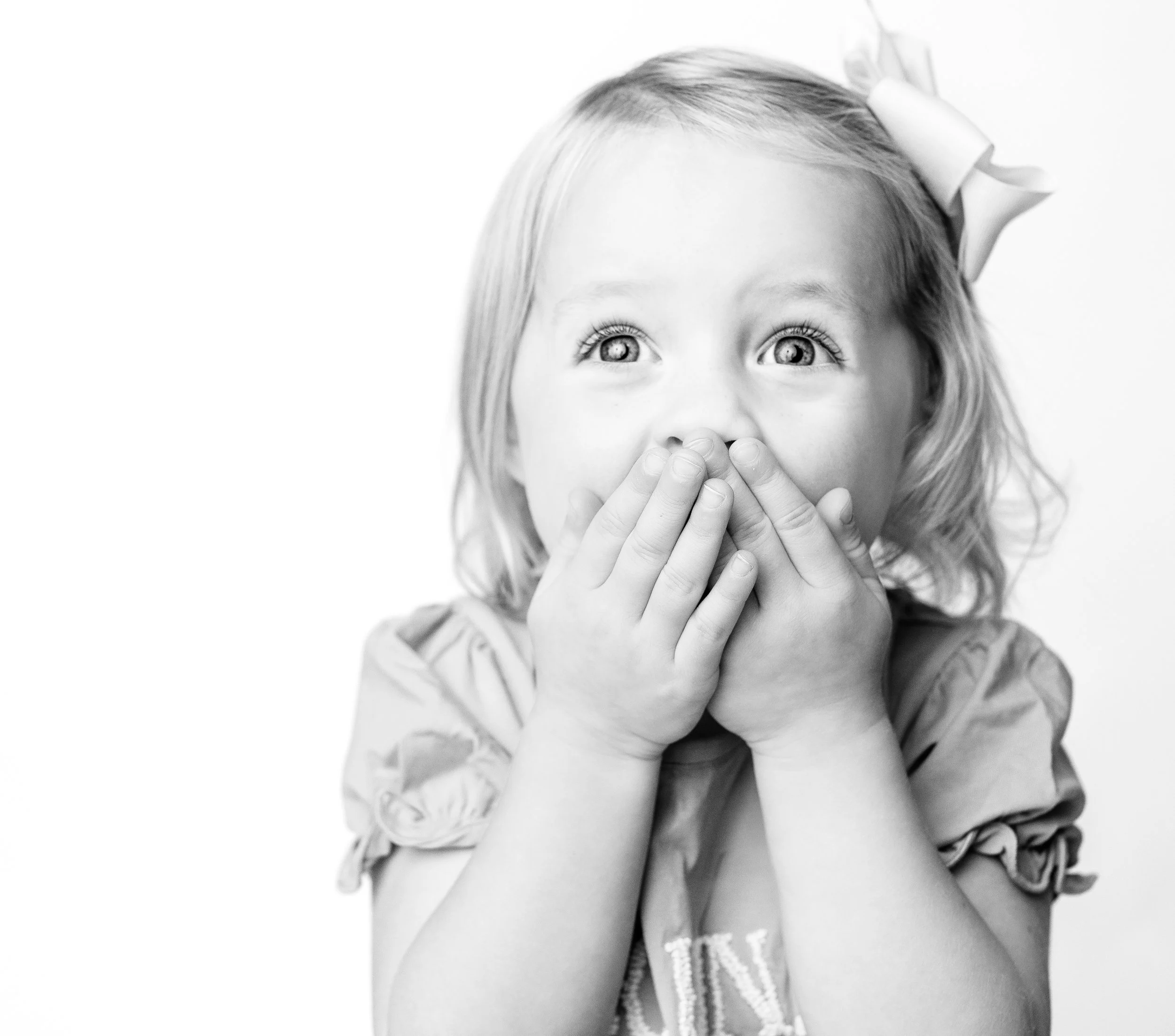 A young girl with light-colored hair, wearing a bow in her hair and a short-sleeved shirt, holds her hands over her mouth with wide eyes, expressing surprise or amazement.