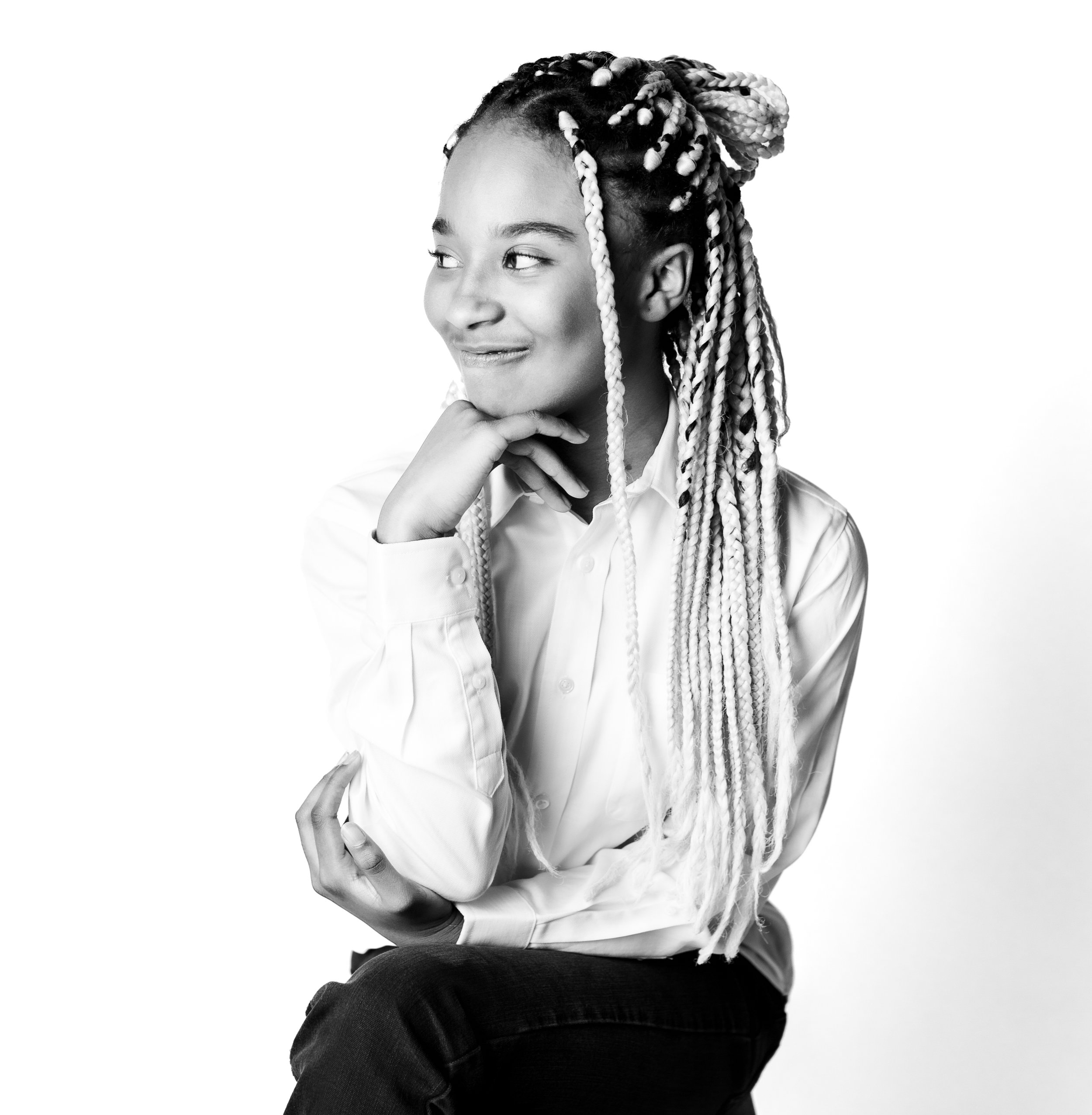 Black and white photo of a woman with long braided hair, wearing a white button-up shirt, sitting and smiling with her hand on her chin against a plain background.