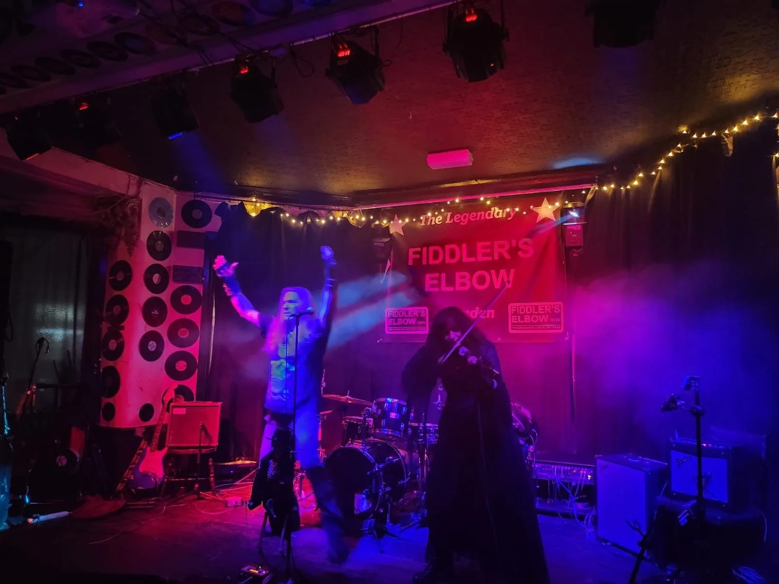 A stage with Symphony of Pain performing at Fiddler's Elbow music venue, illuminated by purple and pink lights, with Pam Chowhan playing violin, and singer Tracie Law with arms raised, all under a sign that reads "The Legendary Fiddler's Elbow".