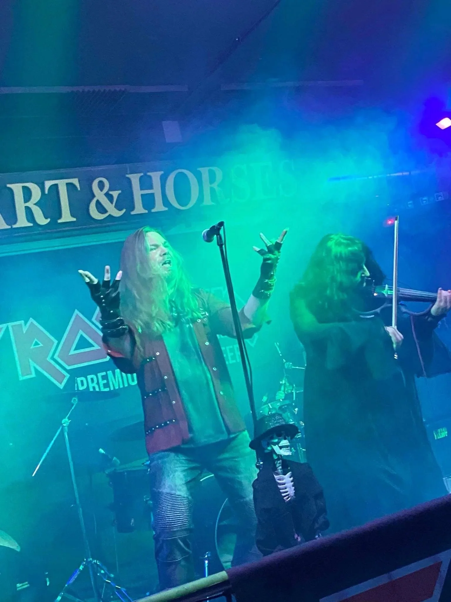 Singer Tracie Law and violinist Pam Chowhan from Symphony of Pain performing on stage at the 'Cart & Horses', which is live music venue, with a sign that reads 'CART & HORSES' in the background, under colorful stage lights.
