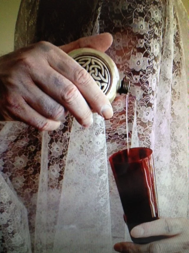 A person's hand holding a watch over a cup of dark liquid, with lace curtains in the background as part of the Symphony of Pain video shoot for Kiss the Bride.