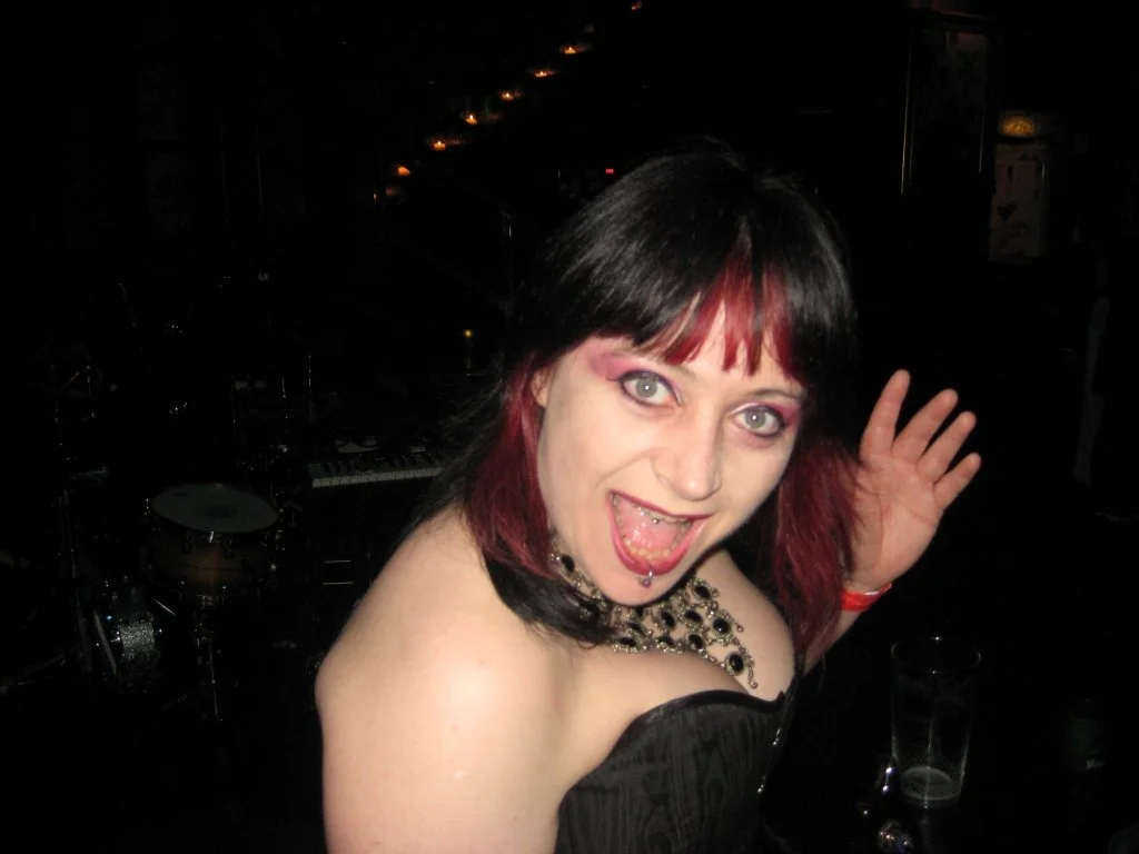 Diane Fox, bassist with Symphony of Pain, with black and red hair posing with her mouth open in a lively indoor setting, with musical instruments and drinks in the background.