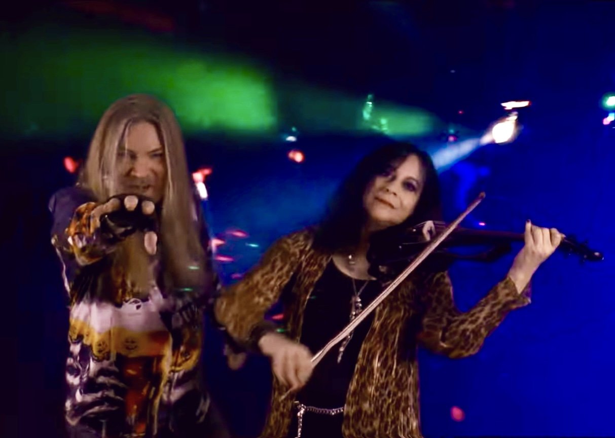 Tracie Law and Pam Chowhan from Symphony of Pain perform on the Barbican main stage, Tracie singing and holding a microphone and Pam playing the violin, with colorful stage lights in the background.