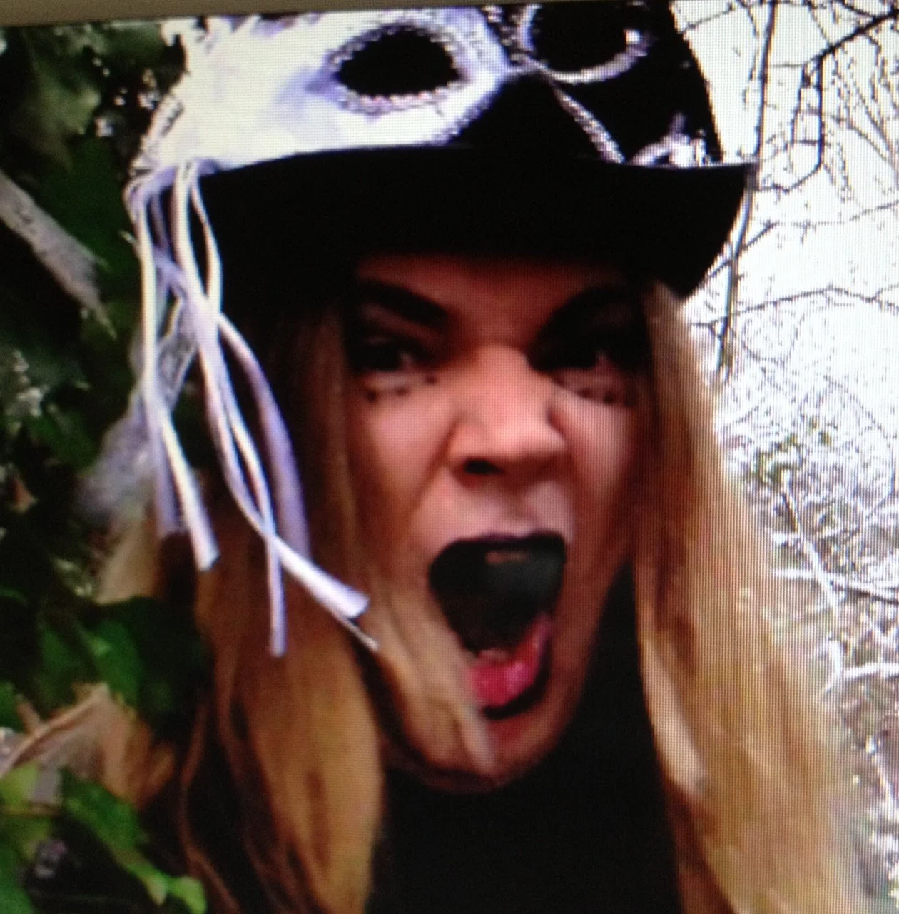 The singer Tracie Law from the band Symphony of Pain wearing a black, white and purple hat and black eyeliner, and has long, blond hair. He is outdoors among trees, with an expressive facial expression, and mouth wide open, singing Kiss the Bride.