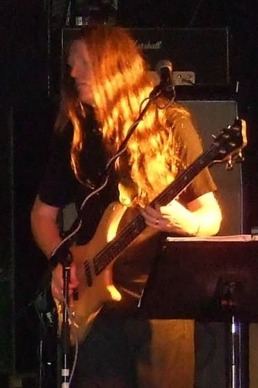 Bassist Mark Bloxsidge from the band Symphony of Pain, with long red hair playing an electric guitar on stage, illuminated by orange stage lighting, with music equipment in the background.