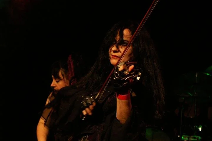 Pam Chowhan, violinist from Symphony of Pain with black hair and a dark outfit playing the violin on stage, with another person partially visible behind her.