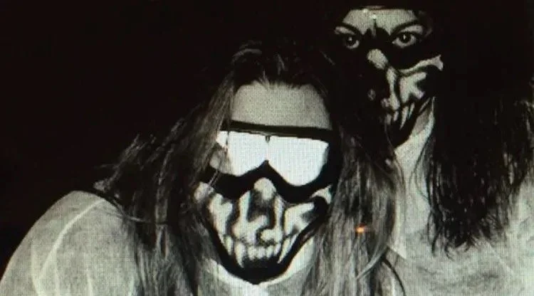 Two people, both in the band Symphony of Pain, wearing masks with skull and horror face designs, one with long dark hair, in a dark setting.