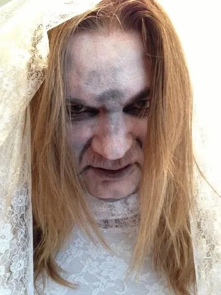 Tracie Law, singer from Symphony of Pain with long blond hair, pale face with dark makeup around eyes, and wearing a wedding veil, looking down with a serious expression. This is from the video Kiss the Bride.