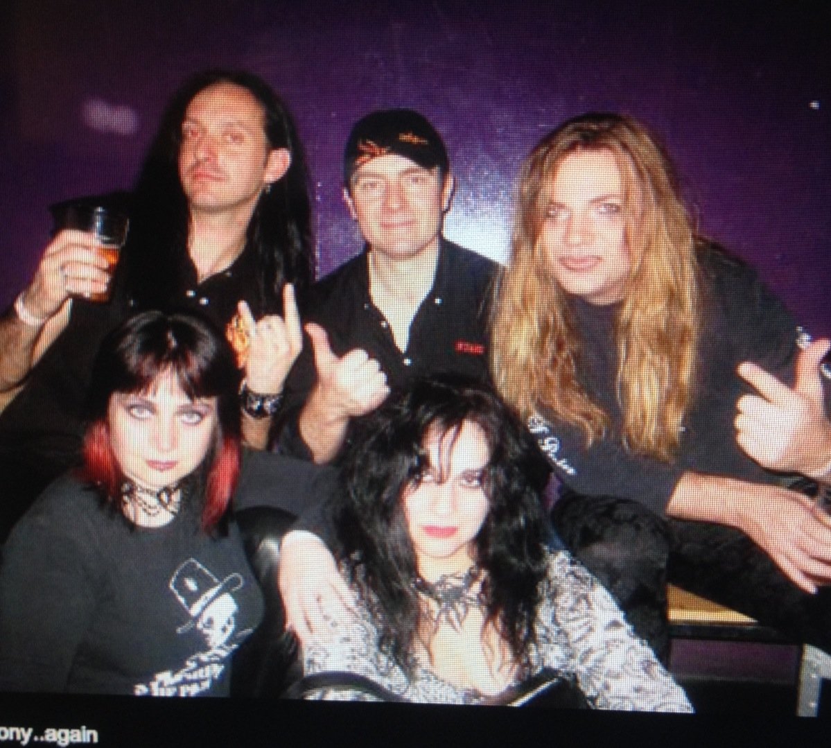 Group of five members of Symphony of Pain with punk and goth style, posing together in a dark setting. Two women are seated in front, while three men stand behind them. All have distinctive hair and makeup, with some giving rock gesture signs.