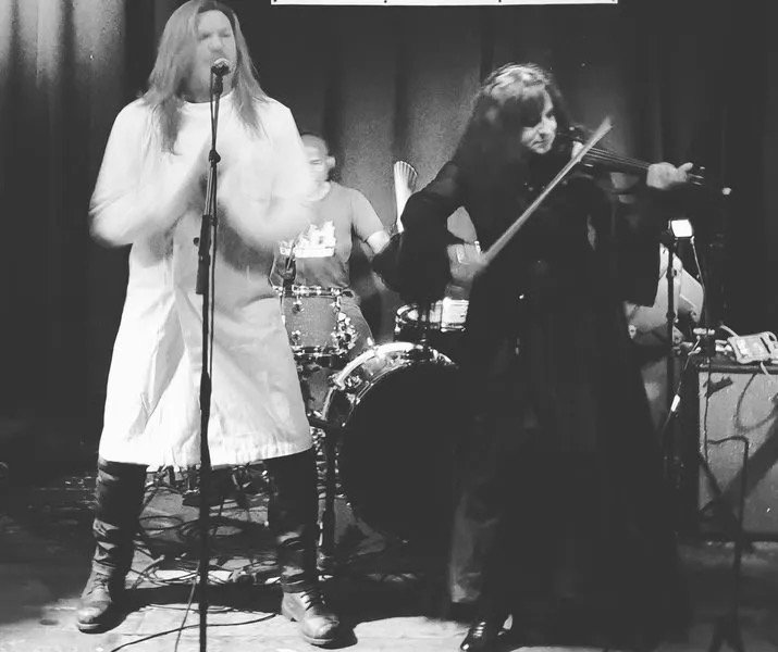 A b&w photo of the band Symphony of Pain performing on stage at the Fiddlers Elbow featuring the singer with long hair and a white coat and the female violinist in a dark outfit, with a drummer and other band members in the background.