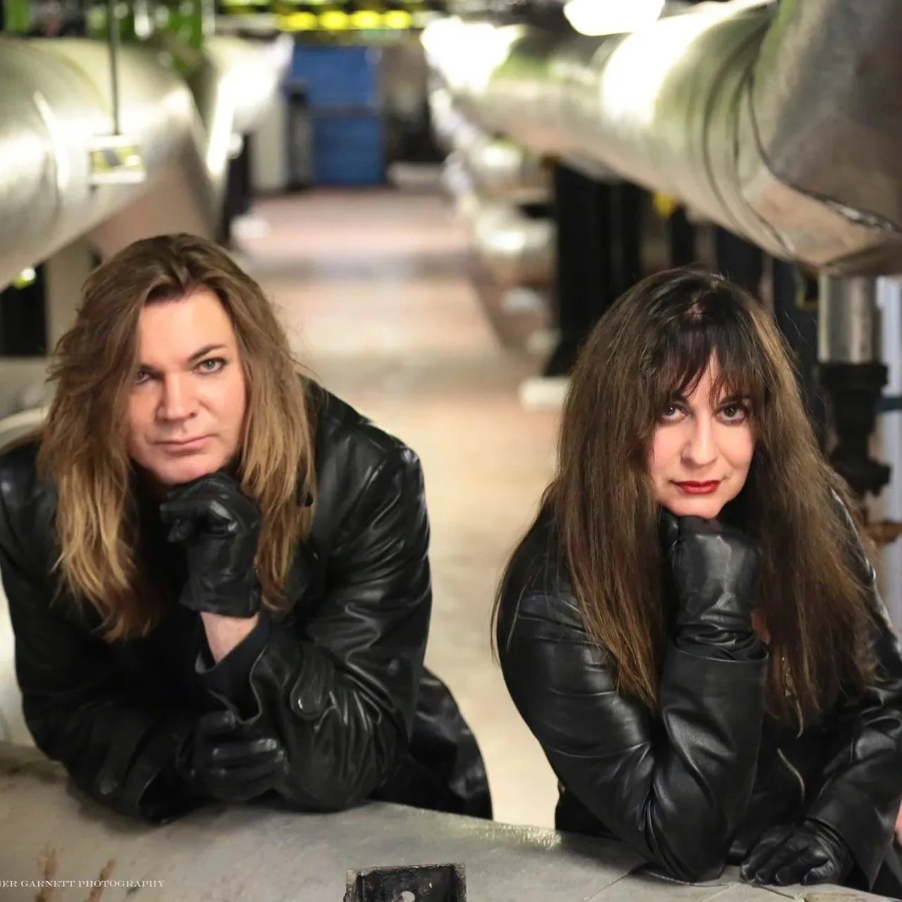 The musicians from the band 'Symphony of Pain', both with long hair wearing black leather jackets and black gloves, resting their chins on their hands, lying on a concrete surface in an industrial setting with large pipes overhead.