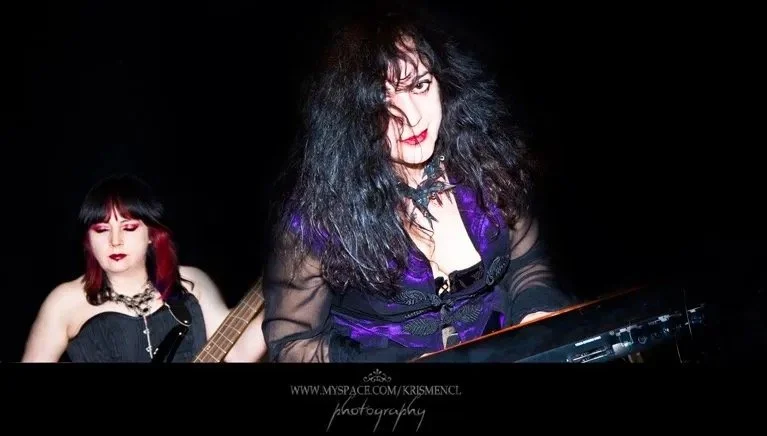 Pam Chowhan playing keyboards and Diane Fox playing bass performing with Symphony of Pain, both dressed in dark clothing with vibrant hair colors.