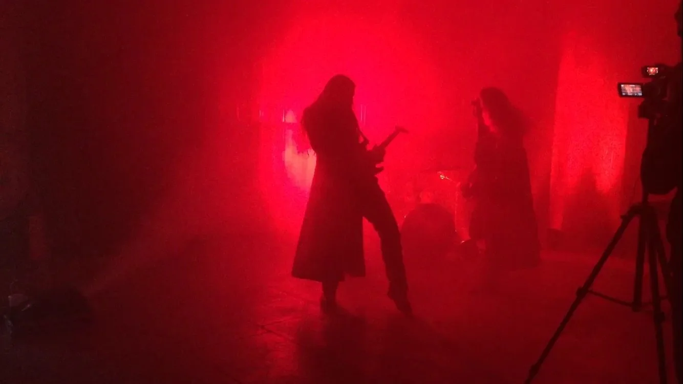 Silhouettes of Symphony of Pain guitarist Jonas Costa and bassist Mark Bloxsidge performing in the video shoot for the track 'Futile' amidst red stage lighting, with a camera filming in the foreground.