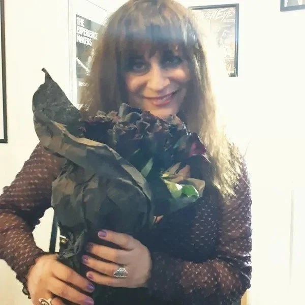 Pam Chowhan, keyboardist and violinist from Symphony of Pain,  with dark curly hair smiling and holding a large bouquet of dark-colored flowers wrapped in black paper.