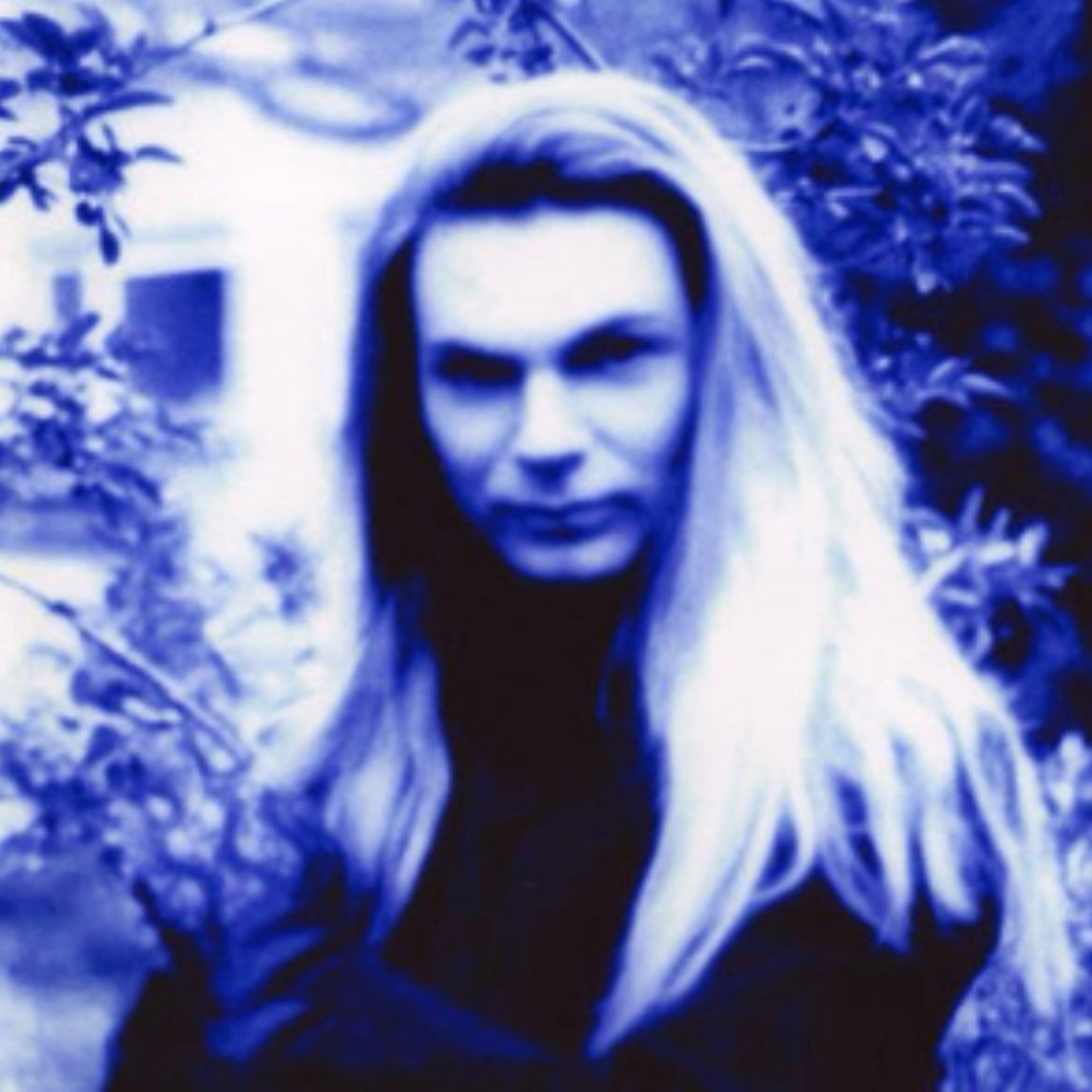 Tracie Law, singer with Symphony of Pain, with long blonde hair outdoors with trees and foliage in the background, the photo is in blue monochrome.