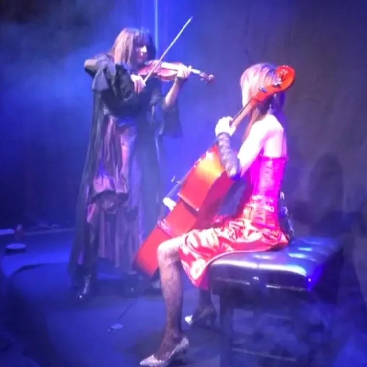 Violinist Pam Chowhan and cellist Jiaxin Lloyd Webber, both in contrasting costumes, on a stage with purple lighting doing a video shoot for the Symphony of Pain track 'Suspicion'.