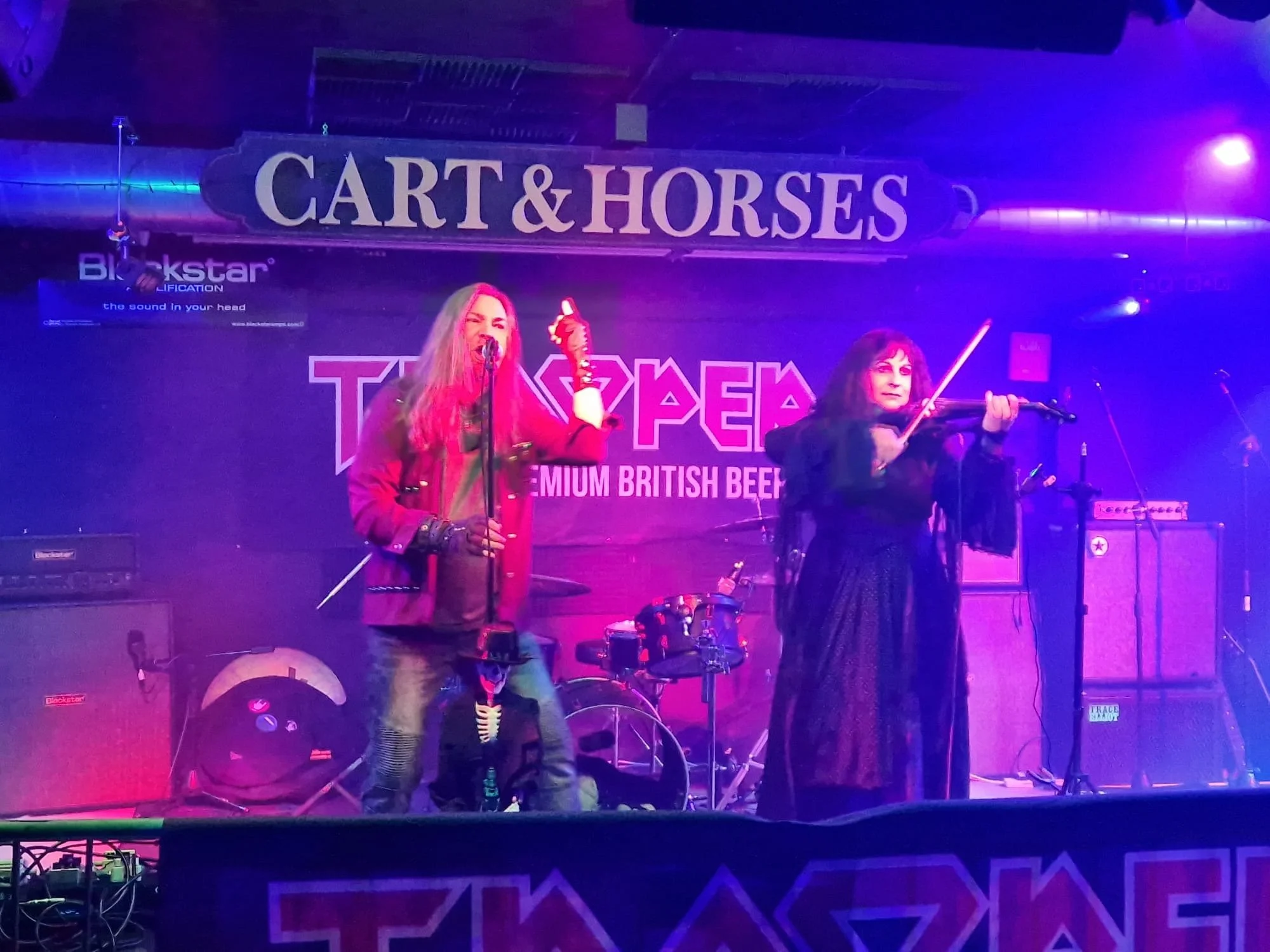 Singer Tracie Law and violinist Pam Chowhan from Symphony of Pain performing on stage at a venue called Cart & Horses, with musical instruments including a violin, drums, and amplifiers visible, under purple and pink lighting.