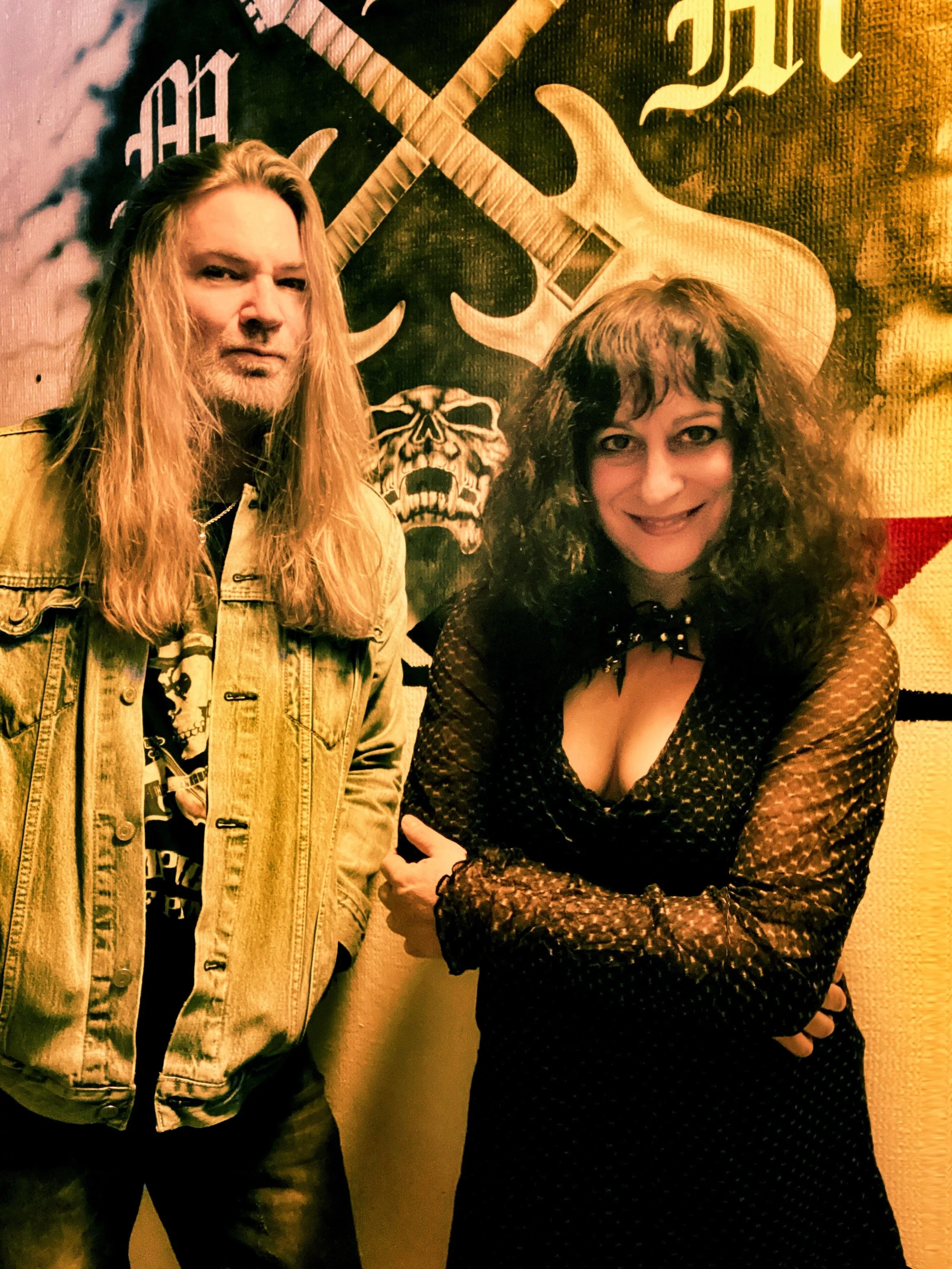 Symphony of Pain band members Tracie Law with long blond hair and a denim jacket standing next to violinist Pam Chowhan who is wearing a purple dress  posing in front of a large banner that features a skull, crossed guitars, and gothic lettering.