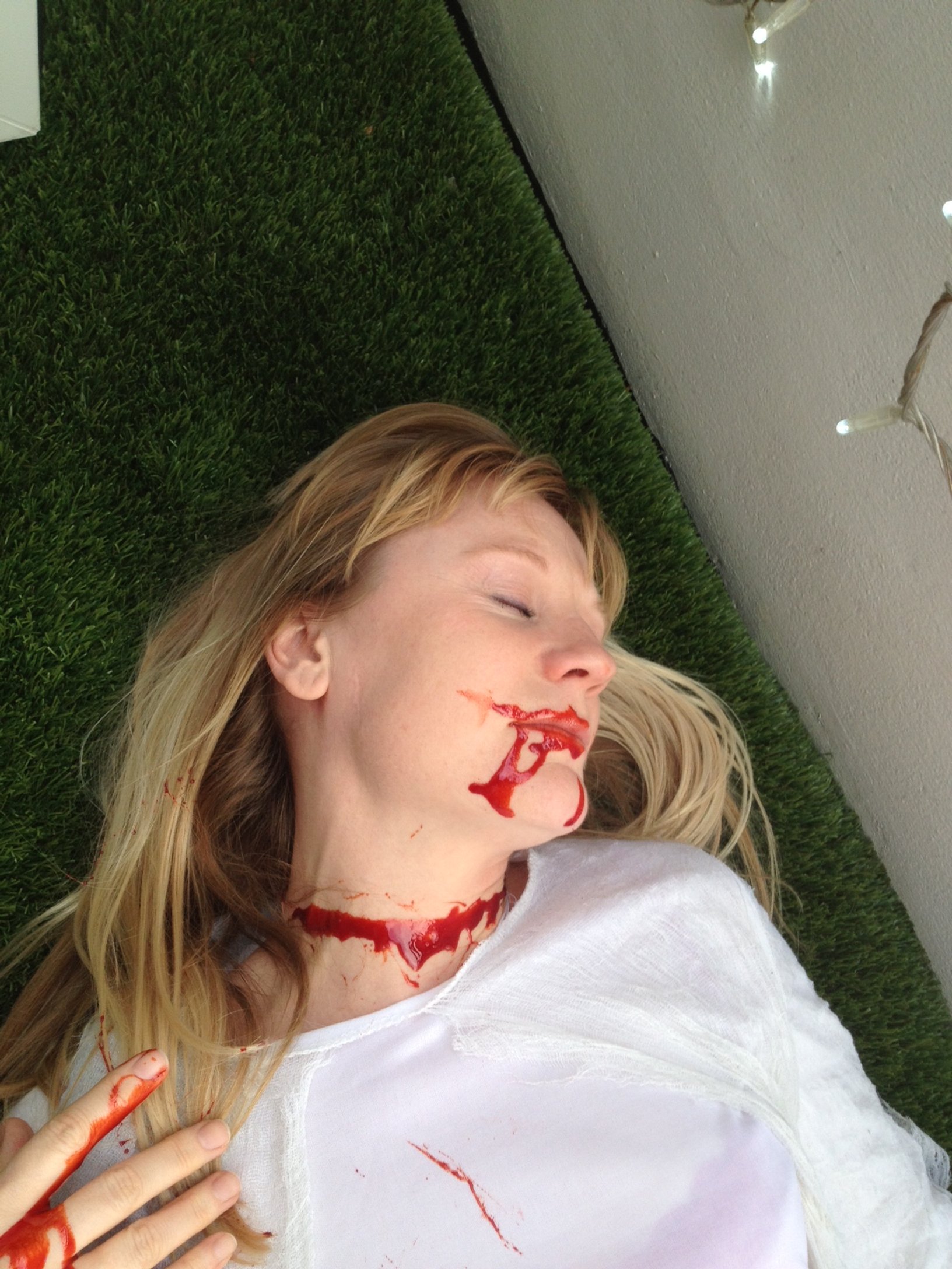 A woman with blonde hair lying on a grassy surface with fake blood on her face, neck, and hand, simulating a horror or Halloween scene as part of a Symphony of Pain photoshoot.