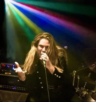 Tracie Law, singer with long blond hair performing on stage with Symphony of Pain with colorful stage lights shining behind him.