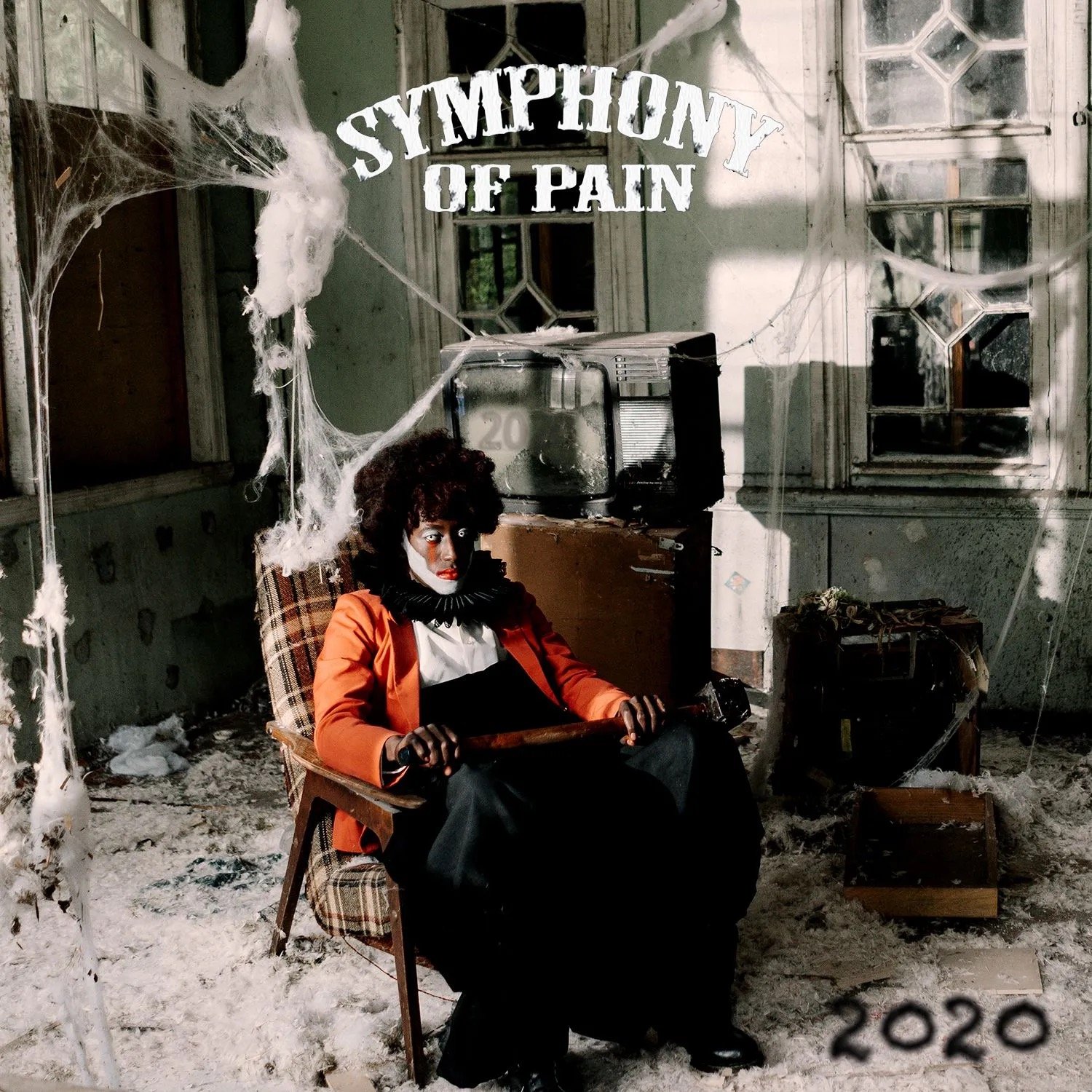 A person dressed as a clown with white face paint, red lipstick, and wearing a black and orange clown costume, sits in a vintage chair inside an abandoned, dusty room decorated with spider webs and cobwebs. The room has old windows and an old televis