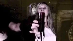 A woman with blonde hair drinking from a glass of dark beverage, with a hand holding the glass.