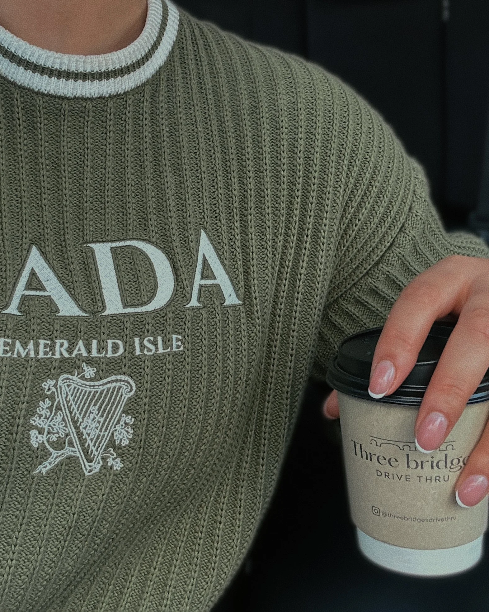 Person wearing a green sweater with 'NAD' and 'Emerald Isle' and a harp logo, holding a coffee cup from Three Bridges Drive Thru.